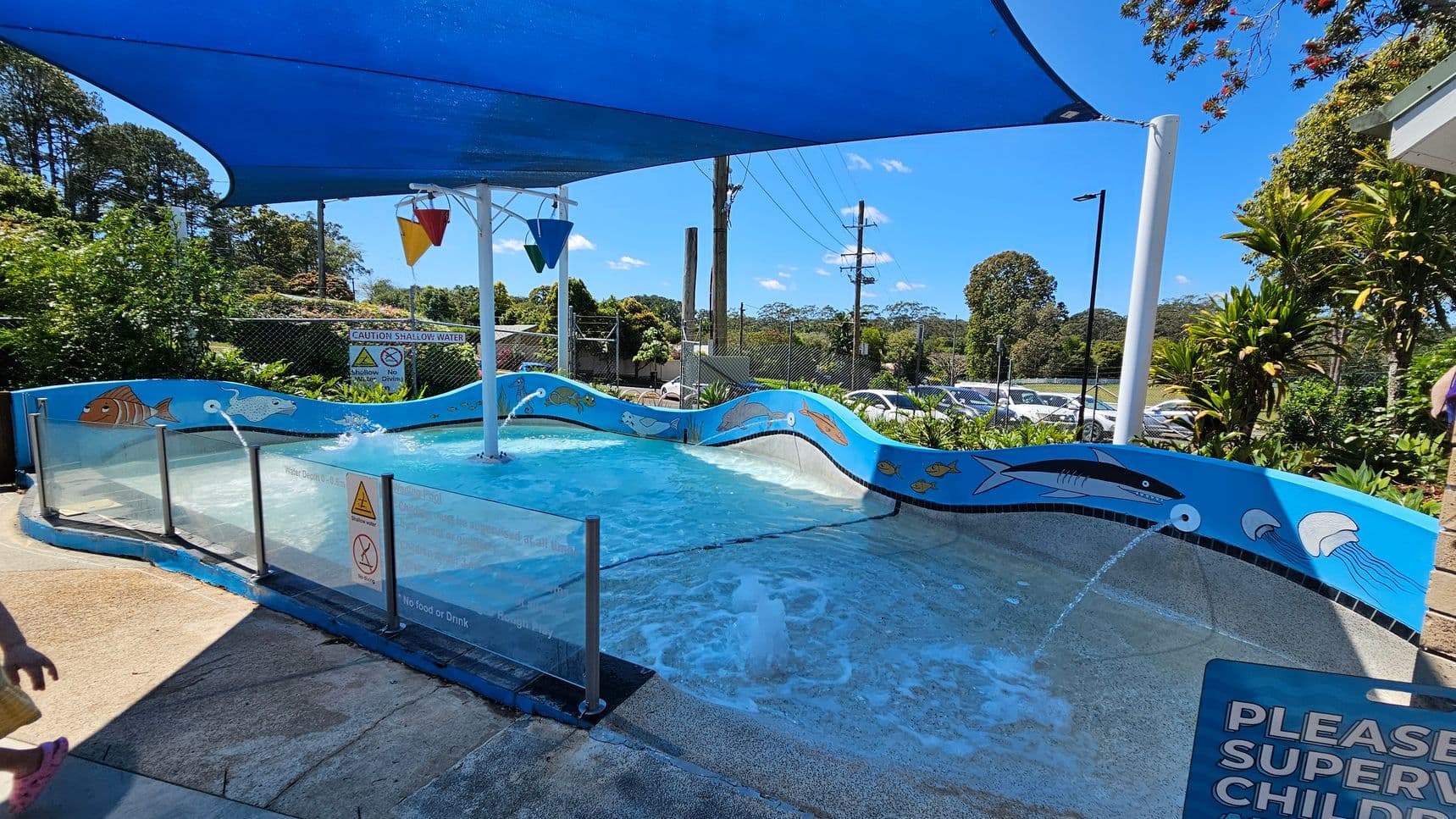 A shallow children's pool with a blue shade canopy overhead. The pool has a wavy blue border decorated with illustrations of sea creatures like fish, dolphins, and jellyfish. In the centre, there is a water feature with colourful buckets that fill and tip over. The pool is surrounded by a glass barrier with caution signs. In the background, there are trees, a fence, and a car park. The foreground shows a concrete floor with a "PLEASE SUPERVISE CHILDREN" sign.