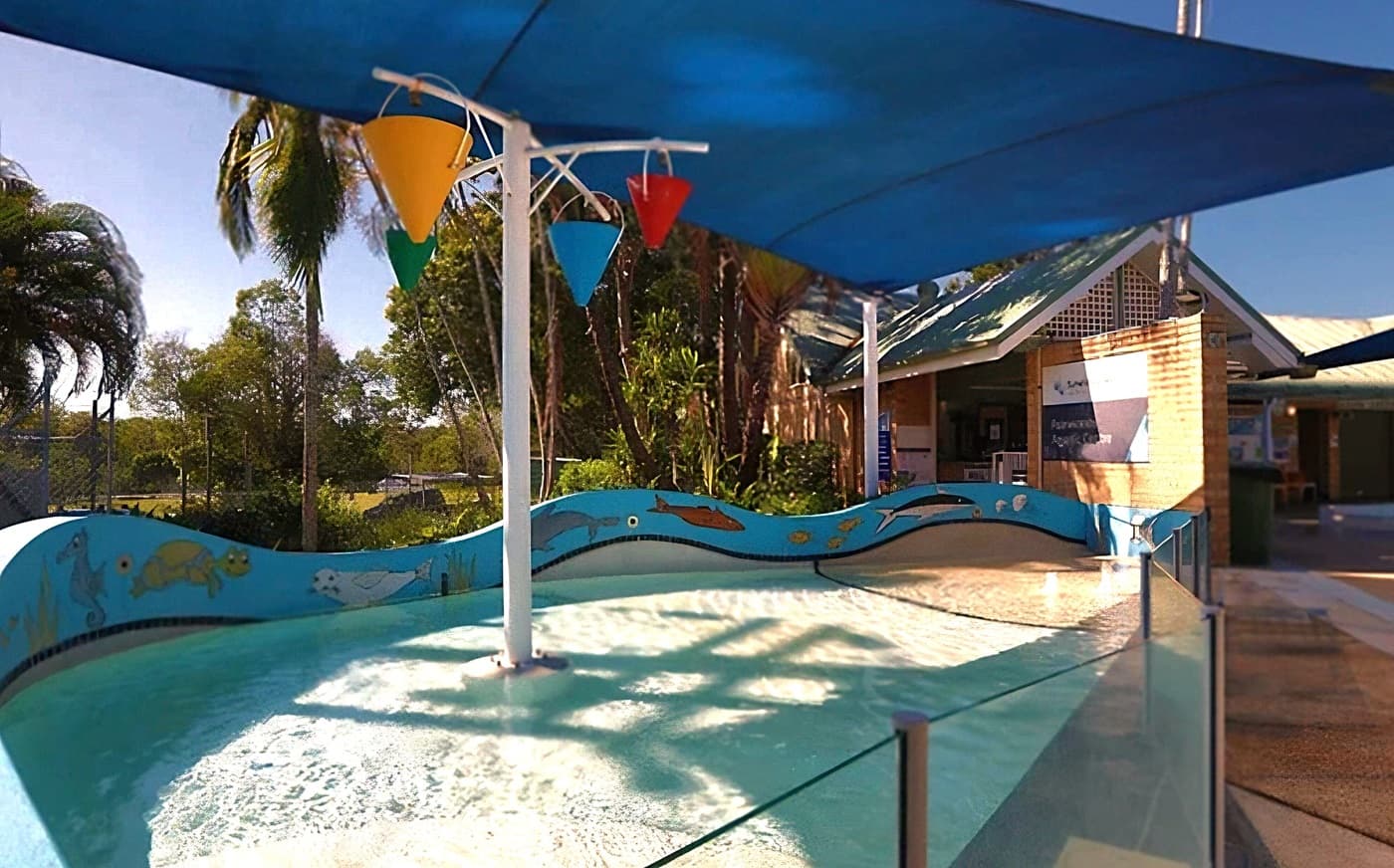 A shallow children's pool with a blue wave-patterned border featuring fish illustrations. In the middle, there is a white pole with three colourful buckets (yellow, green, and red) that pour water. A blue shade sail is overhead. On the right, a brick building with a sign reading "Sunshine Coast Palmwoods Aquatic Centre" and a few people near the entrance. In the background, a chain-link fence, trees, and parked cars.