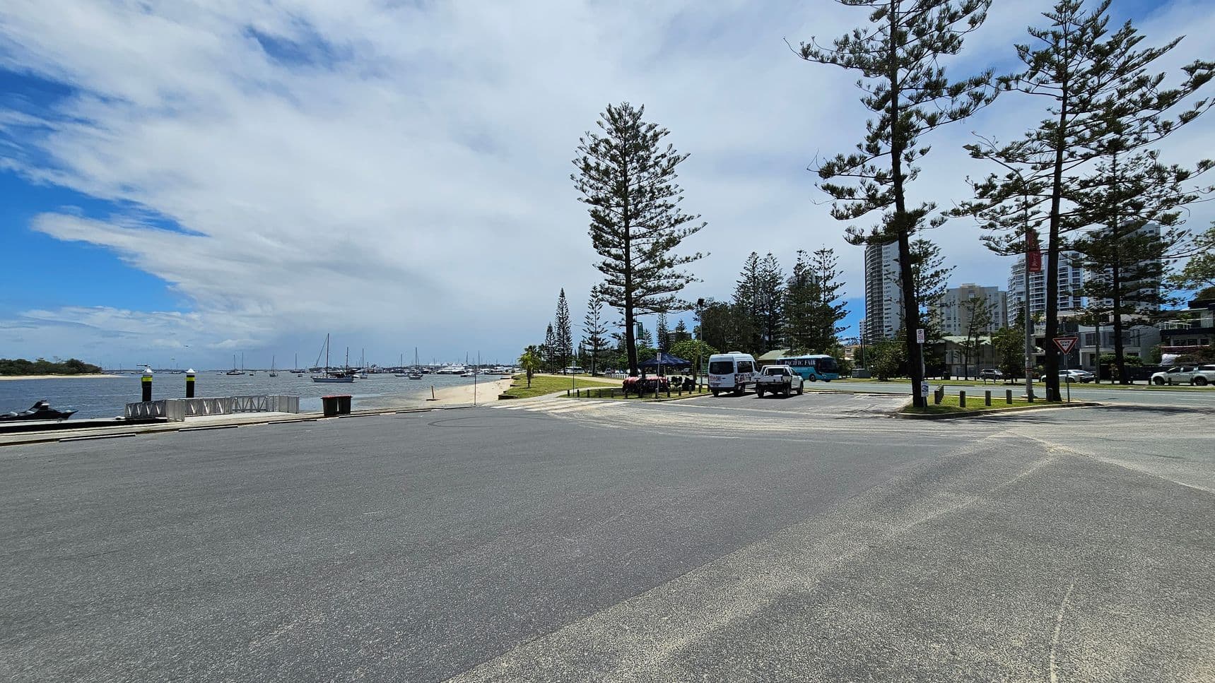 A coastal scene with a wide road in the foreground. On the left, a body of water with several moored sailboats and a small sandy beach. In the middle, tall pine trees line the grassy area, with a few parked vehicles, including a van and a bus. On the right, modern high-rise buildings are visible behind more trees. The sky is partly cloudy, with patches of blue visible.