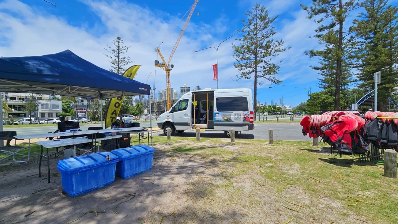 On the left, a blue canopy tent covers tables with various items and two large blue storage bins. In the centre, a white van with its side door open is parked on a grassy area. To the right, a rack holds numerous red life jackets. In the background, there are tall trees, a construction crane, and buildings. The ground is a mix of grass and sand, and a road with parked cars runs behind the setup.