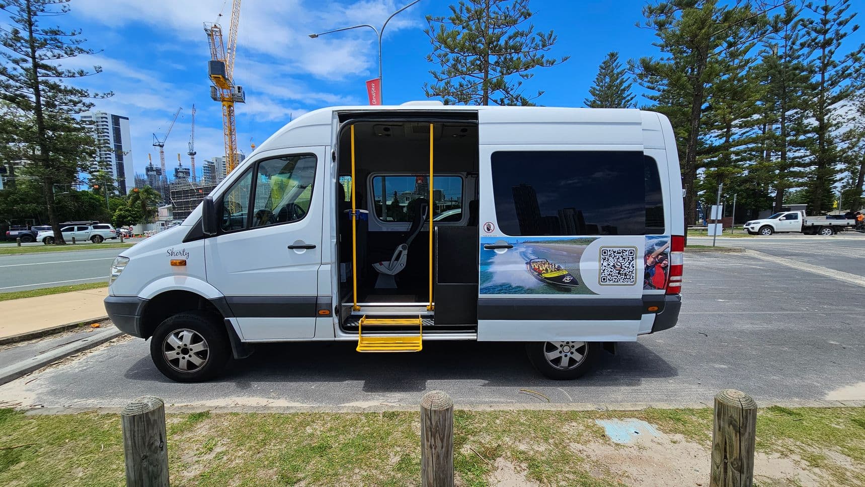 A white van with an open sliding door and yellow steps is parked on a paved area. The van has a graphic of a speedboat and a QR code on its side. In the background, there are tall pine trees and several construction cranes near high-rise buildings. A few vehicles are visible on the road, and the ground is a mix of grass and sand.