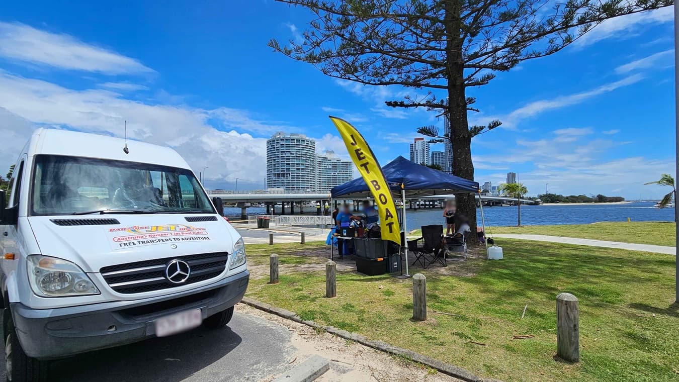 A white van with a Mercedes logo is parked on the left, featuring signage for jet boat rides. On the right, a yellow flag with "JET BOAT" written on it stands next to a blue canopy tent. Under the tent, people are gathered around a table. In the background, a river is visible with a bridge and modern buildings. The area is grassy with a few trees, and wooden posts line the path.