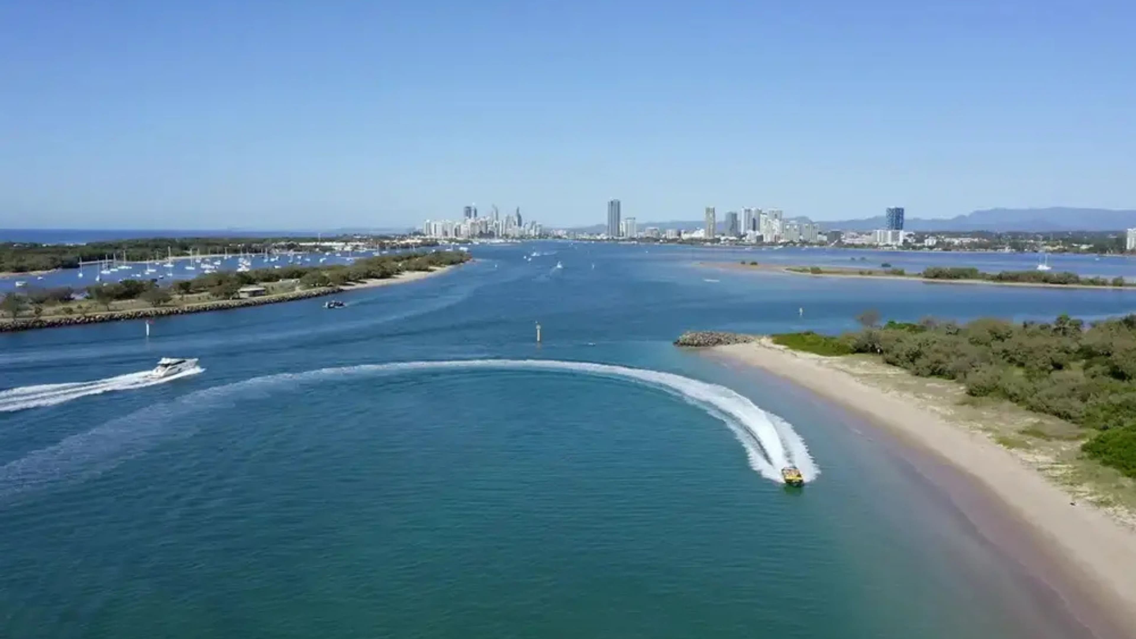 A yellow speedboat with passengers is moving swiftly along the water near a sandy beach. The boat creates a large spray of water as it speeds by. On the left, the water is a deep blue, while on the right, the sandy beach stretches out with some greenery and trees in the background. A rocky breakwater is visible in the distance on the left side.