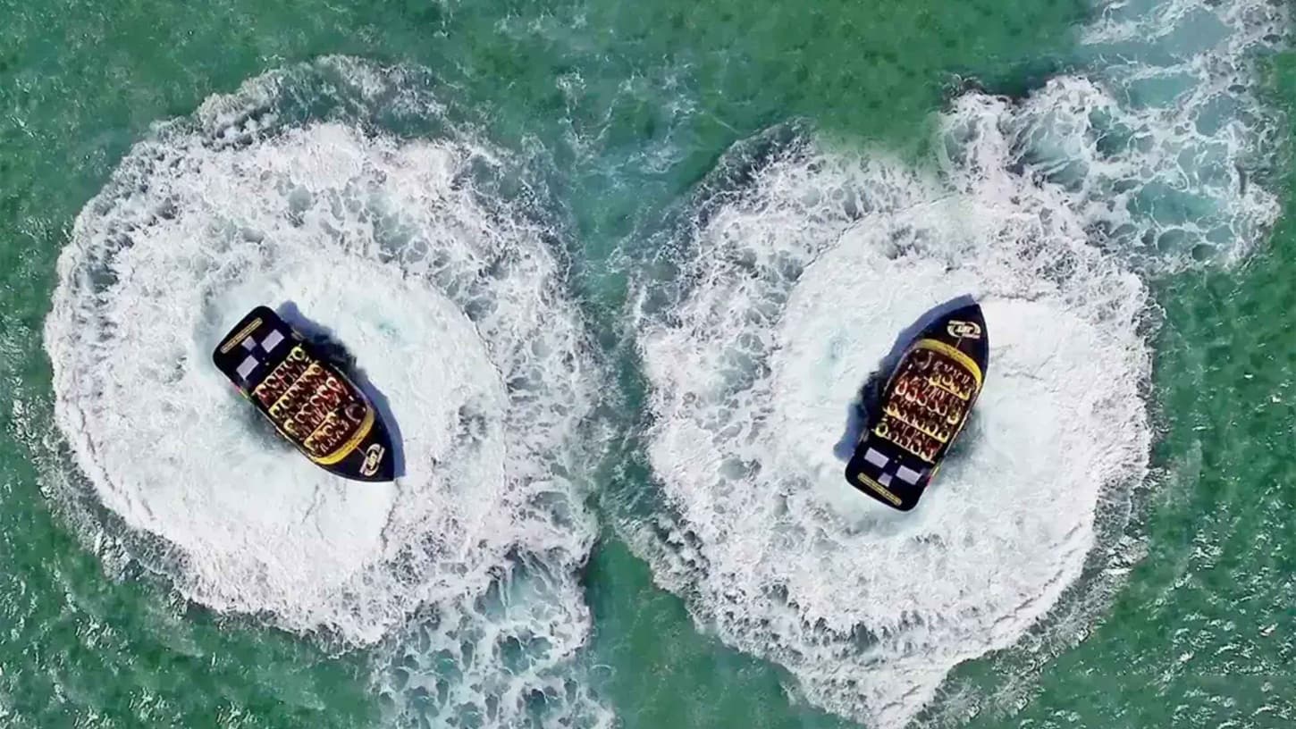 Two speedboats are captured from above, each creating circular patterns in the water as they spin. The boats are black with yellow accents and are filled with passengers wearing life jackets. The water is a clear turquoise, and the wake from the boats forms distinct white swirls around them. The scene is dynamic, with the boats positioned side by side, each in the centre of its own circular wake.