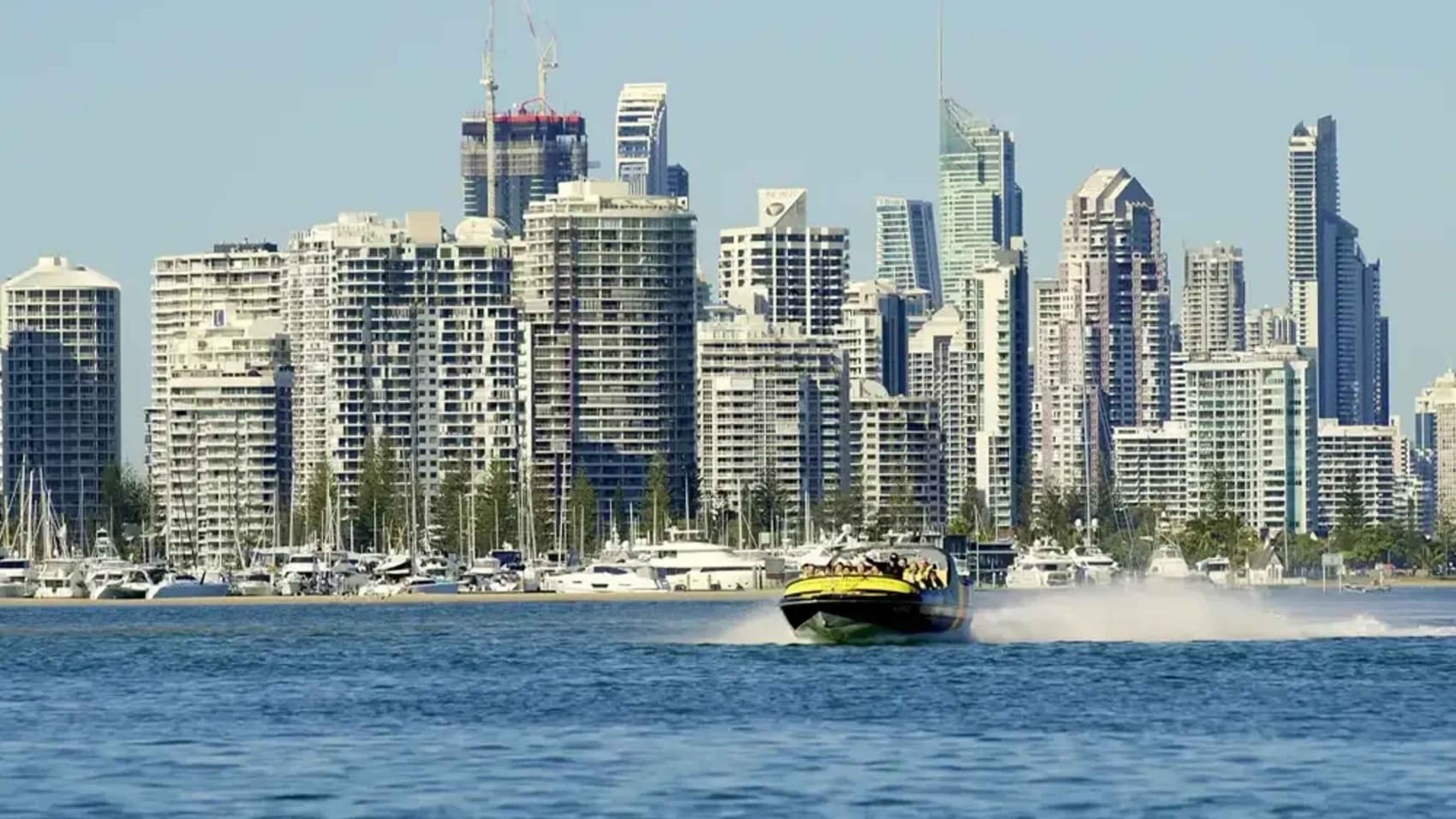 A speedboat moves swiftly across the water in the foreground, creating a trail of spray behind it. In the background, a dense cluster of modern skyscrapers and high-rise buildings form a city skyline. Numerous yachts and boats are docked along the waterfront, with masts visible. The water is a deep blue, contrasting with the light-coloured buildings. The sky is clear, providing a bright backdrop to the scene.