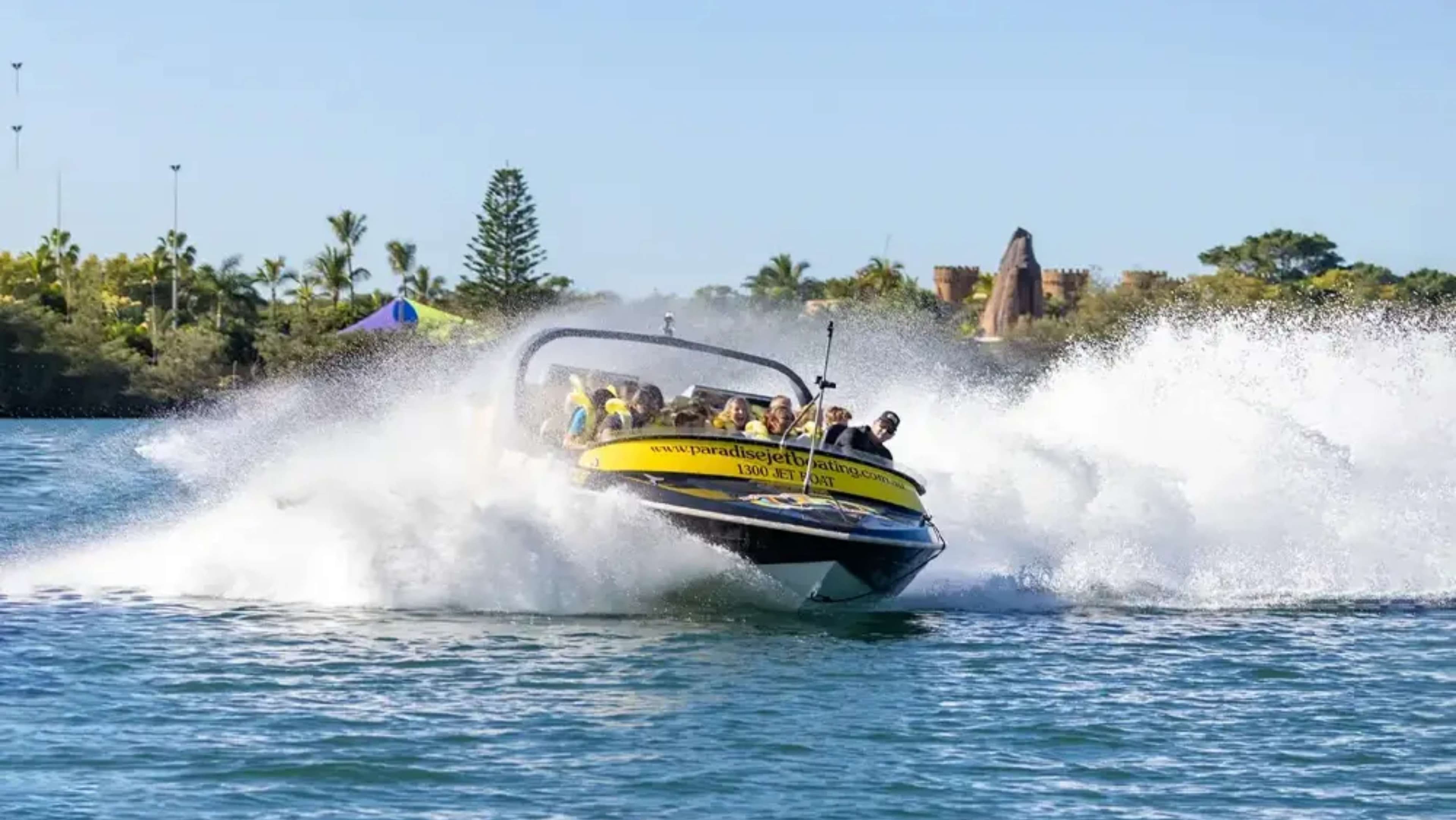 On the left, there are palm trees and a vibrant tent with a blue top, suggesting a recreational area. In the foreground, a jet boat is cutting through the water, creating splashes as it accelerates. The boat has a yellow and black colour scheme, with several passengers visibly enjoying the ride. To the right, there are more trees and what seems to be a decorative stone structure or castle-like feature in the background, possibly part of a theme park.