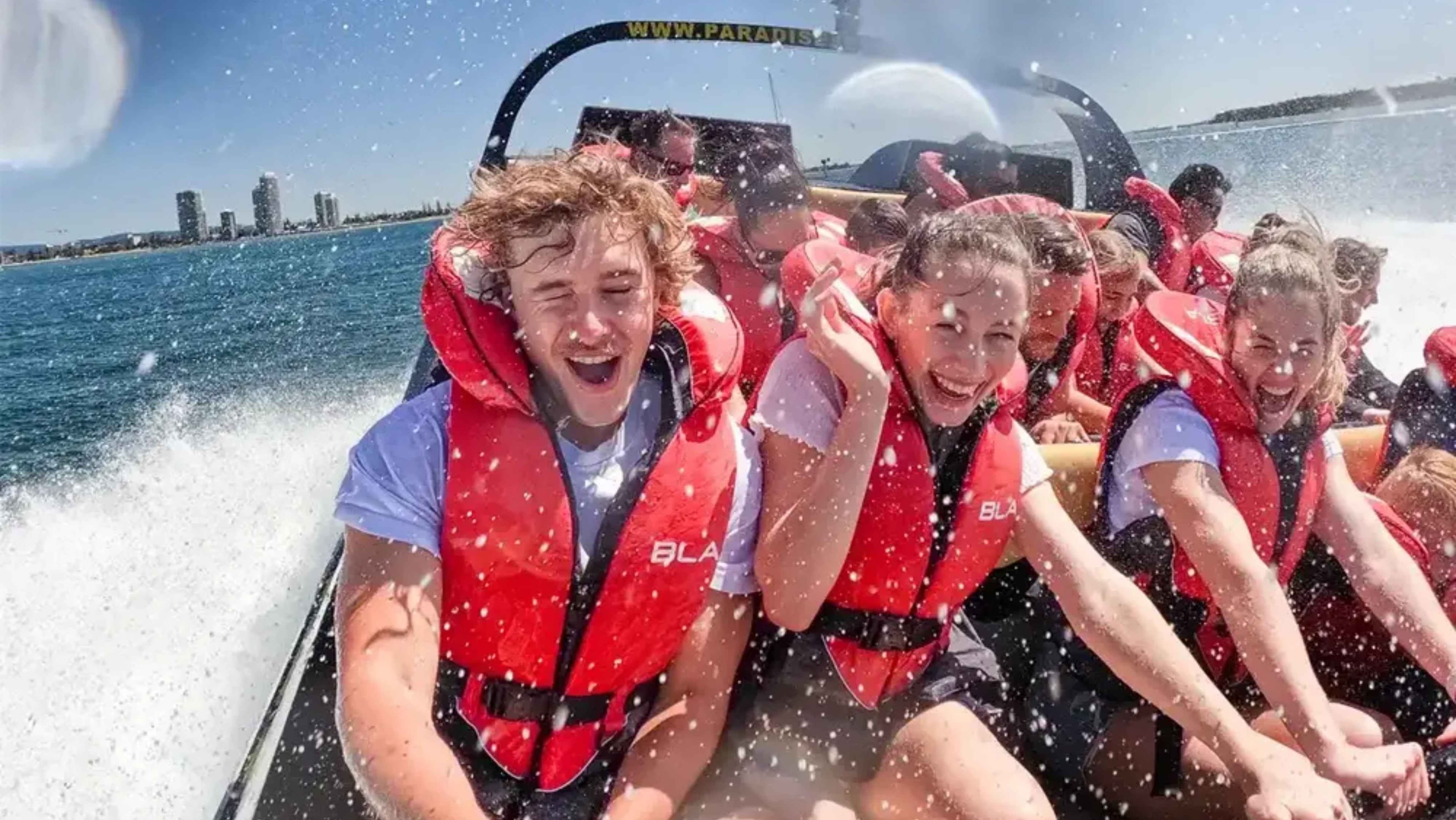 A group of people wearing red life jackets are seated on a speedboat, gripping the safety bars. Water splashes around them, indicating high speed. The background shows a city skyline with several tall buildings against a clear blue sky. The boat's wake is visible on the left, creating a dynamic scene. The people appear to be enjoying the ride, with expressions of excitement.