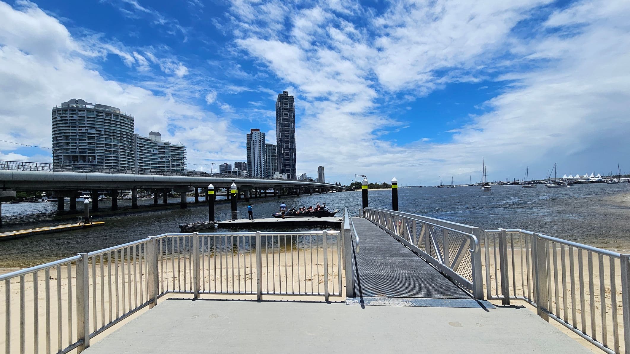A sandy beach is on the left, with a dock extending into the water. A small boat is moored at the dock. In the background, several sailboats are visible on the water. On the right, there is a paved area with parking spaces and a few tall trees. Beyond the trees, modern high-rise buildings are visible. The scene is calm, with a mix of natural and urban elements.