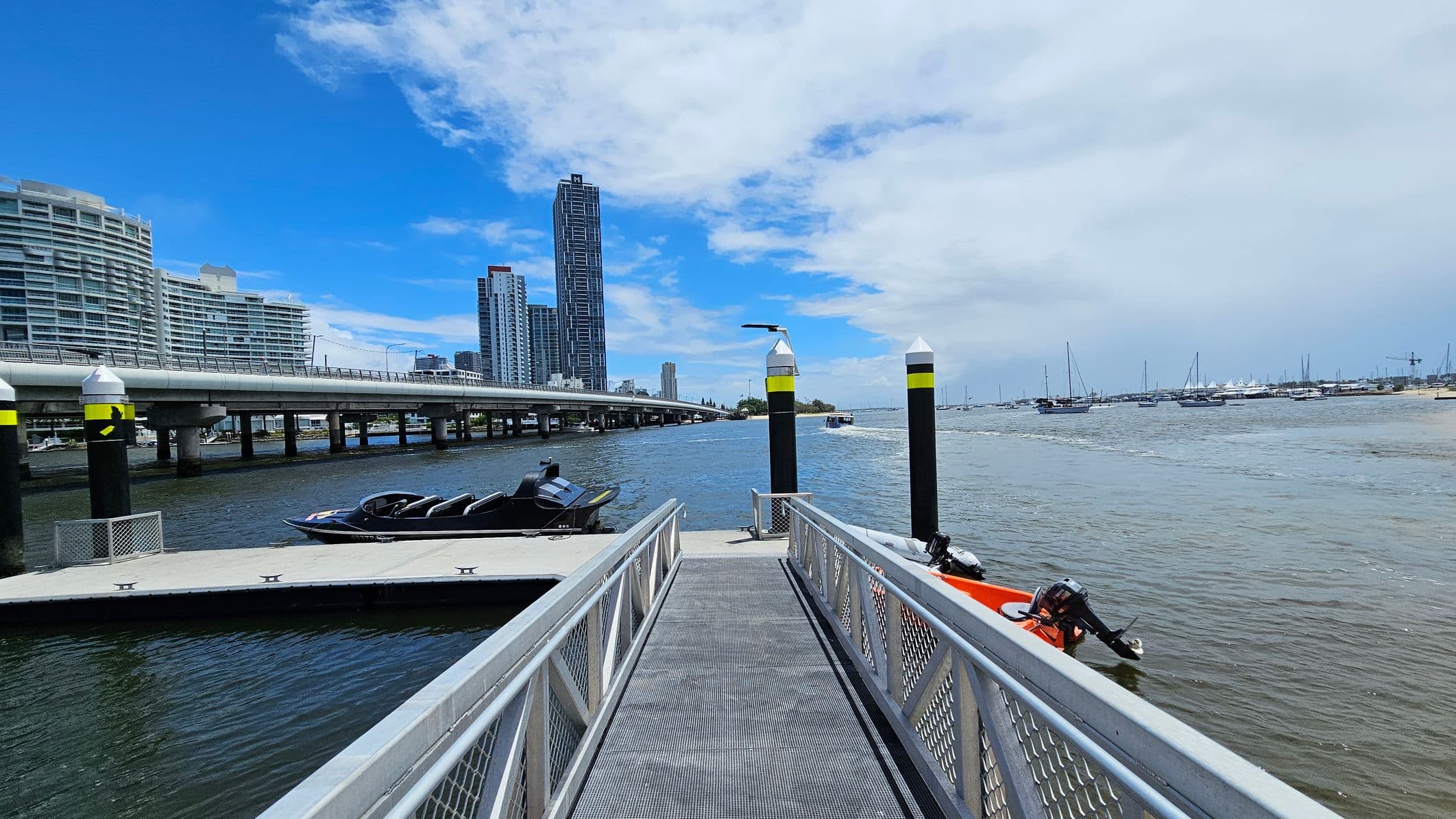 A metal walkway leads to a floating dock on a body of water. On the left, a sleek black boat is moored. In the background, a bridge stretches across the water, with modern high-rise buildings visible beyond it. On the right, several sailboats are anchored in the distance. The water is calm, and the sky is partly cloudy. Two black and yellow posts are positioned near the dock.