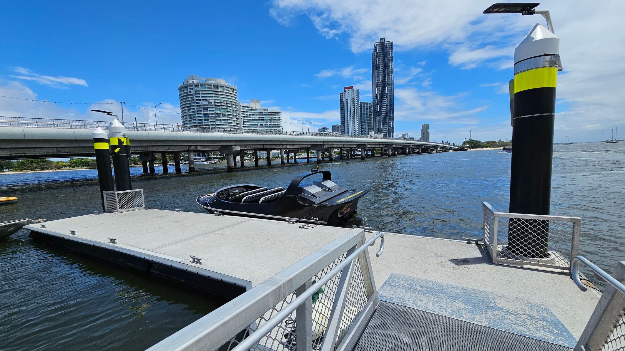 A modern dock with a sleek black boat moored on the right. The dock is bordered by metal railings and has a textured surface. Two tall black and yellow posts with solar panels are positioned on the dock. In the background, a bridge spans across the water, with several high-rise buildings visible, including a cylindrical one on the left and a tall rectangular one on the right. The water is calm, and a few boats are seen in the distance.