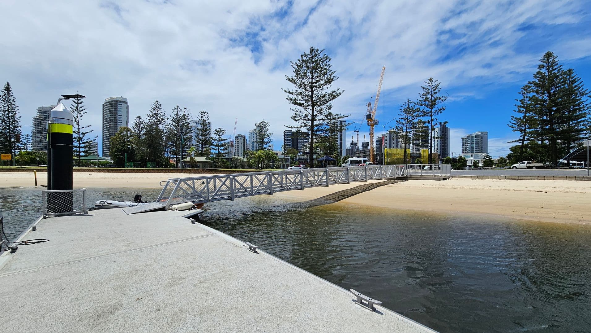 A concrete dock extends into the water on the left, with a black and yellow post at the edge. A metal walkway leads from the dock to a sandy beach on the right. In the background, tall buildings and construction cranes are visible, surrounded by tall pine trees. A road runs parallel to the beach, with a few vehicles on it. The sky is partly cloudy.