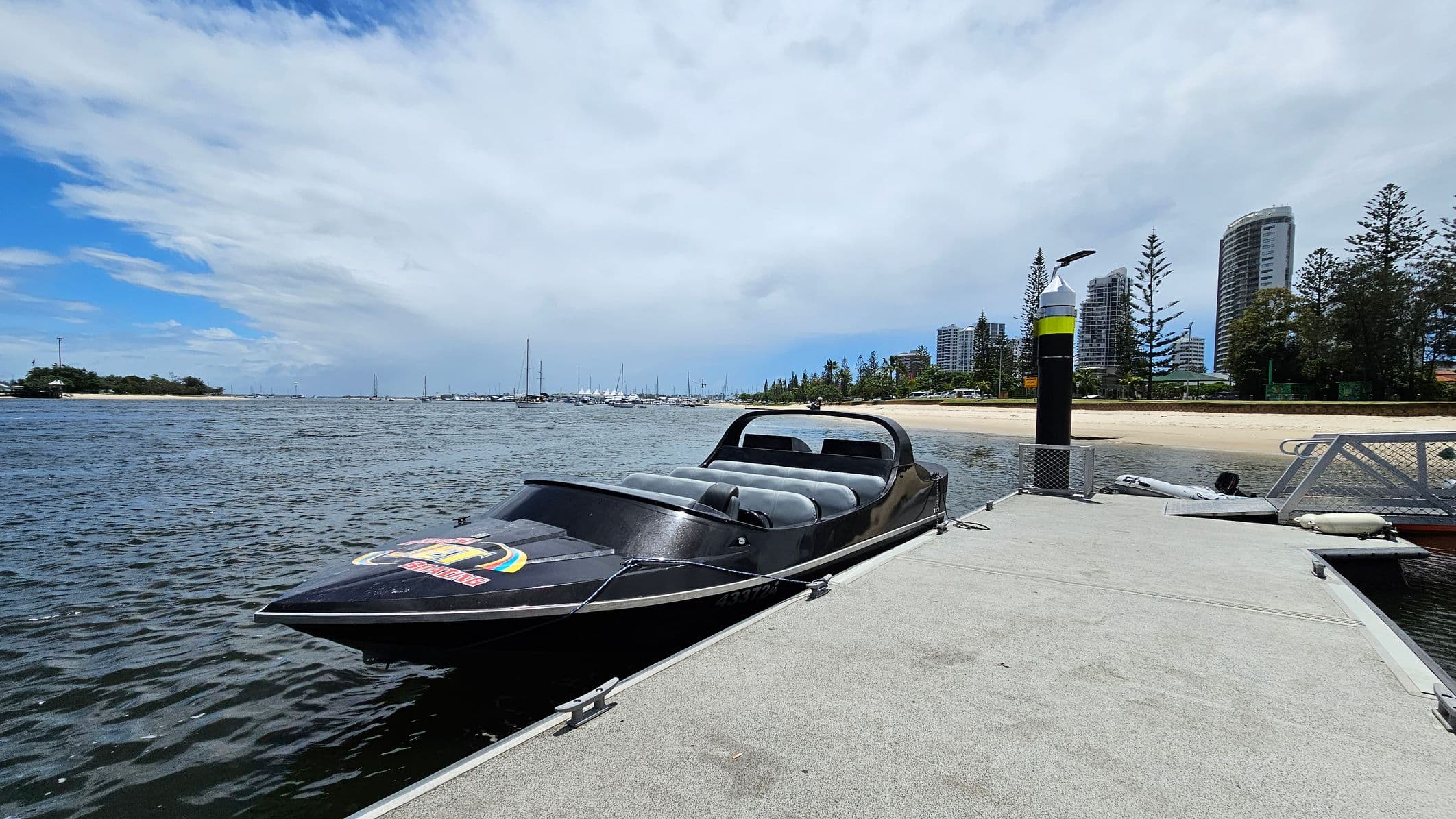 A sleek black speedboat is moored to a concrete dock on the right. The boat has a low profile with grey seating. In the background, a sandy beach is lined with tall pine trees and several modern high-rise buildings. On the left, the water stretches out with numerous sailboats anchored in the distance. A black and yellow striped post is visible on the dock.