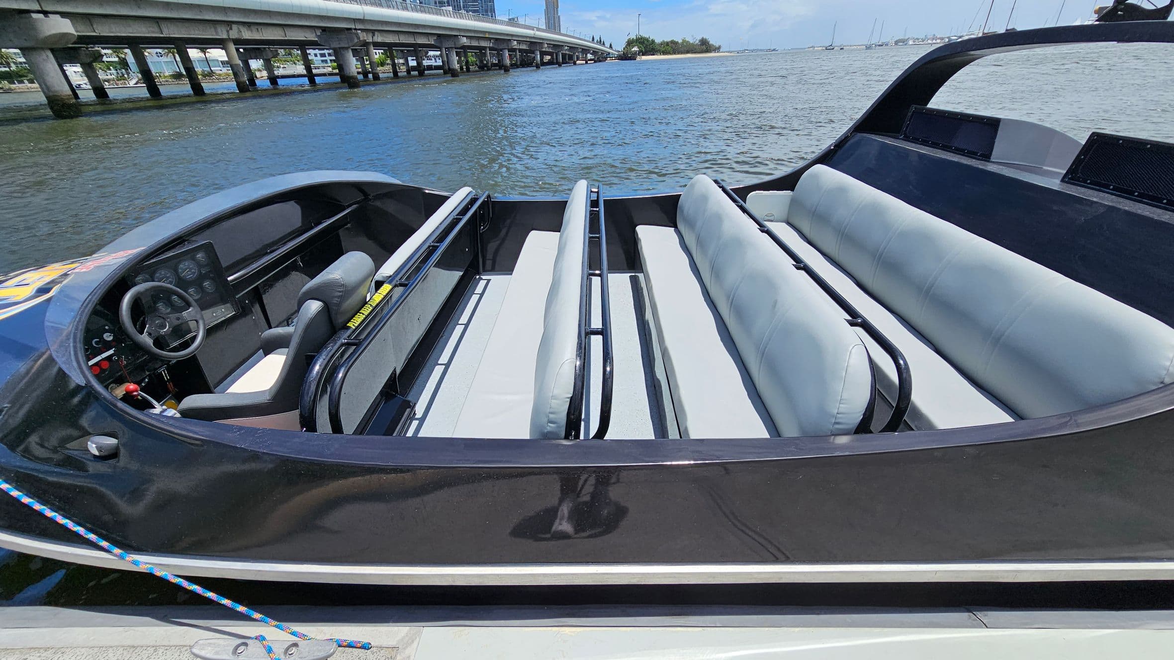 A sleek black speedboat with a modern design is docked by the water. The interior features two rows of light grey cushioned seats with black railings. The cockpit area has a steering wheel and a control panel with various gauges and buttons. A colourful rope is tied to the boat's cleat. In the background, a bridge extends over the water, with buildings and trees visible in the distance. The water is calm, and several boats are seen further away.