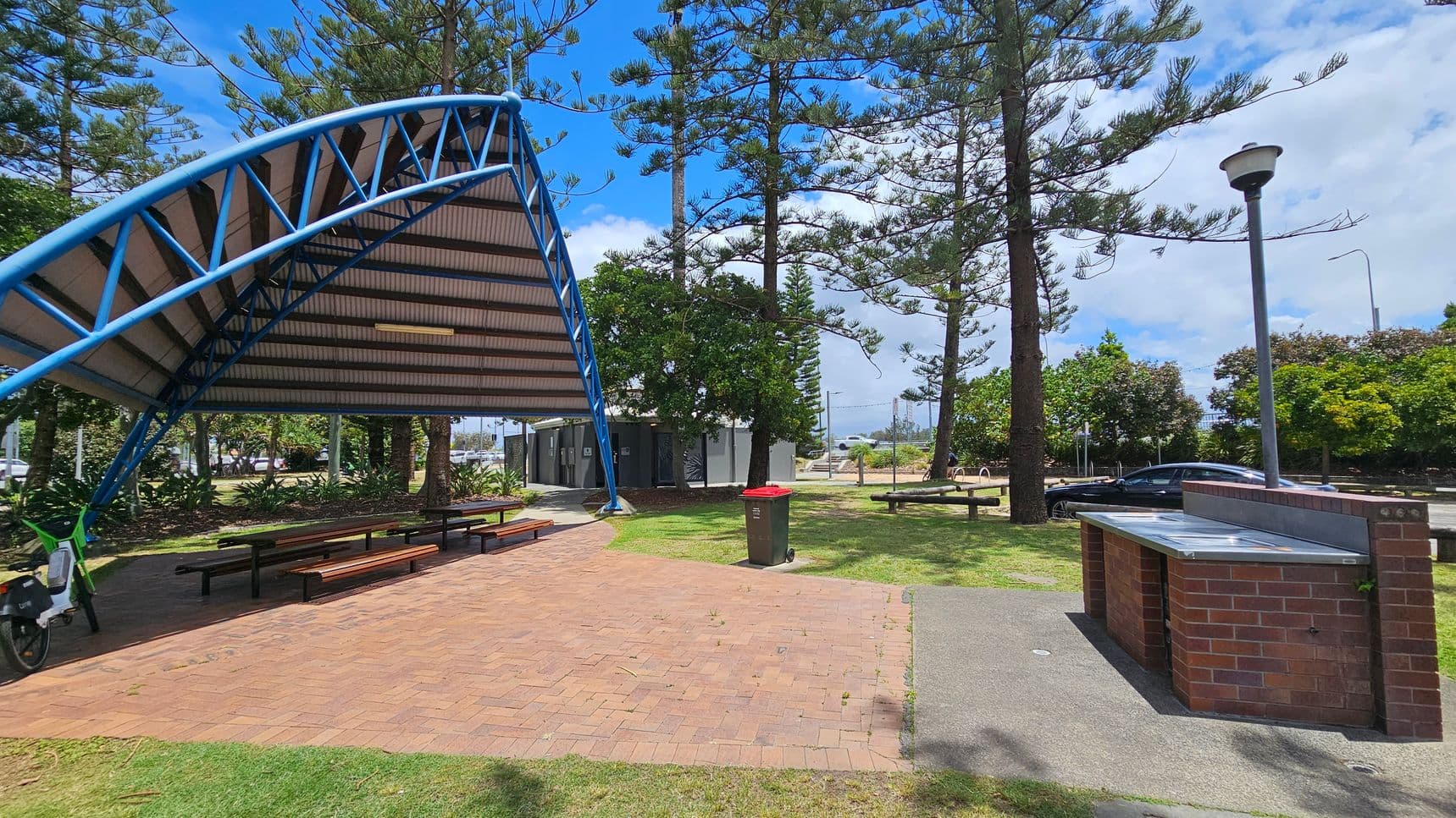 A park scene with a covered picnic area on the left, featuring a blue metal frame and a corrugated roof. Beneath the shelter are several picnic tables. A bicycle is parked nearby. In the centre, there is a brick path leading to a bin with a red lid. On the right, a brick barbecue stand is visible, along with a lamp post. Tall trees and bushes are scattered throughout the grassy area, and a car is parked in the background.