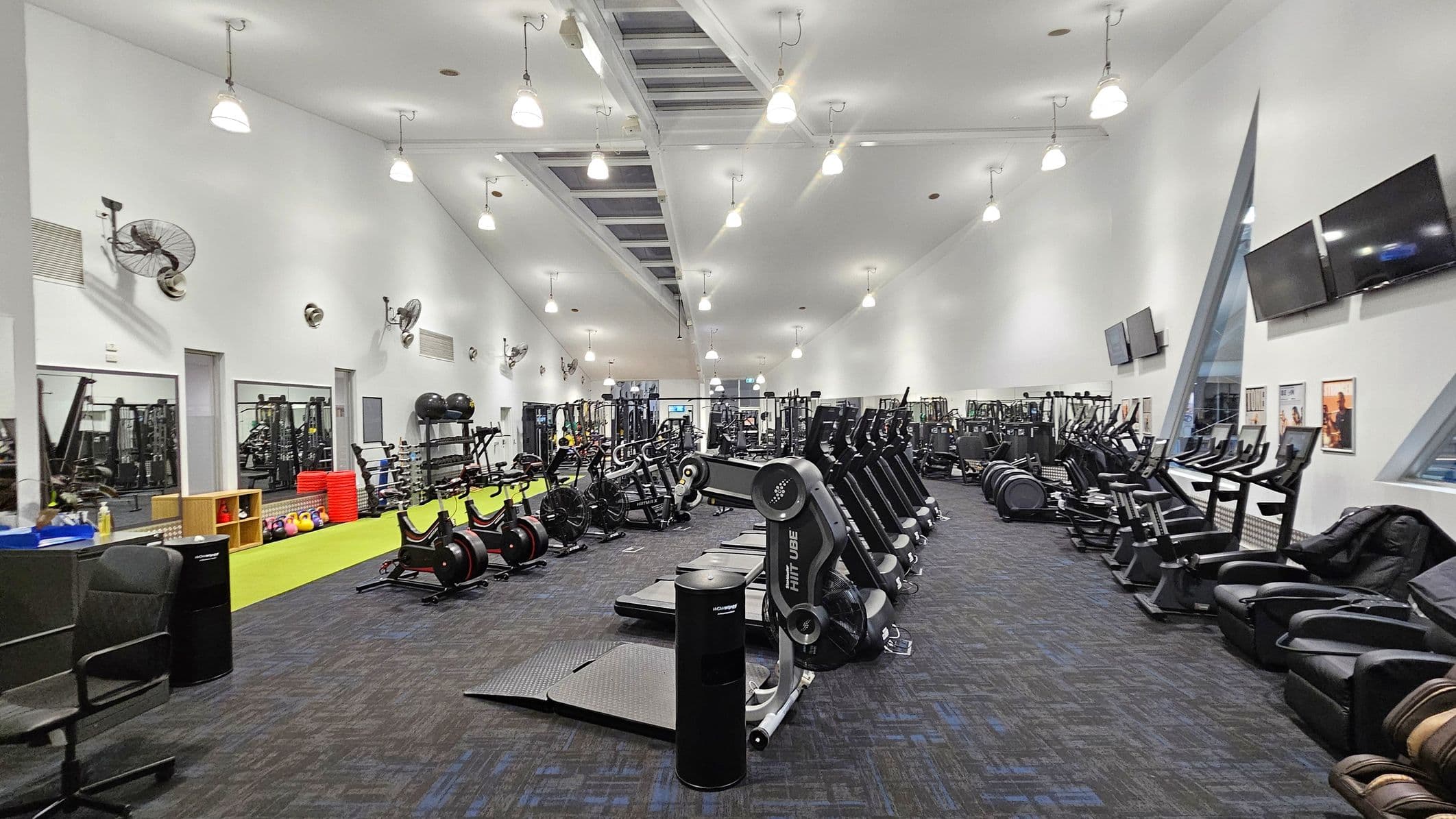 A spacious gym interior with a high ceiling and bright lighting. On the left, there are wall-mounted fans, mirrors, and a variety of gym equipment including weights and exercise balls. In the centre, rows of exercise machines such as treadmills and stationary bikes are neatly arranged. On the right, there are large windows, wall-mounted televisions, and more exercise machines. The floor is carpeted in dark tones, and the ceiling features hanging lights.
