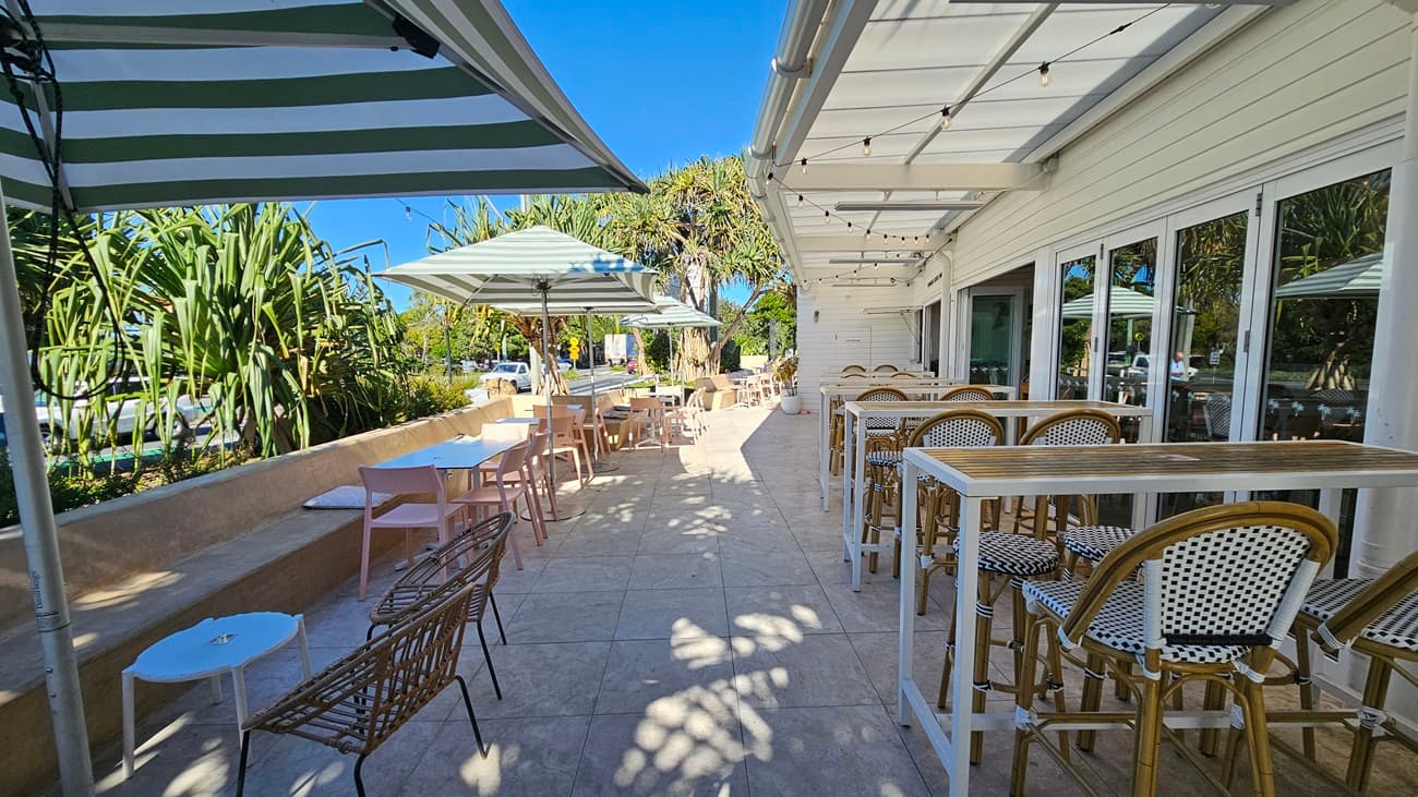 On the left, there are green plants and a beige stone bench with a small white table and a wicker chair. In the middle, there are several tables and chairs under green and white striped umbrellas on a tiled floor. On the right, there is a white wooden wall with large windows and high tables with bar stools. The background includes more plants and trees, with a glimpse of the road. The ceiling has string lights hanging above the seating area.