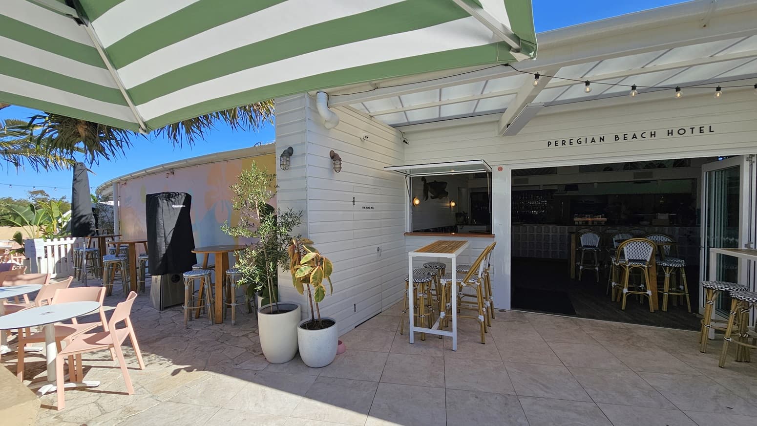 On the left, there are pink chairs and white tables under a green and white striped umbrella, with potted plants nearby. In the middle, there is a white wooden wall with a small bar table and stools, leading to the entrance of the Peregian Beach Hotel. On the right, the entrance has more bar tables and stools inside, with a tiled floor extending throughout. The ceiling is a white pergola with beams, providing partial shade. The background includes more plants and part of the hotel building.