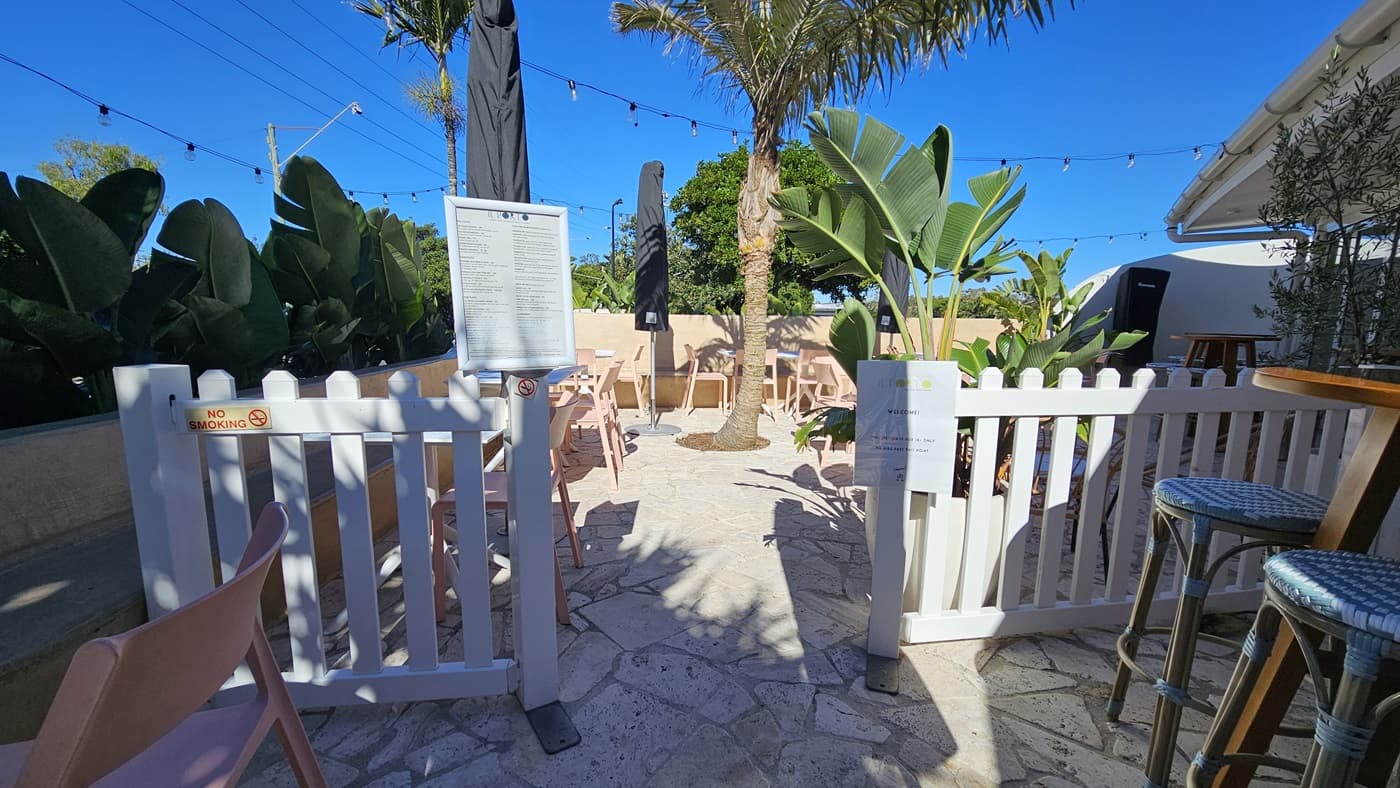 On the left, there are pink chairs and tables next to a white picket fence, with large green plants behind them. In the middle, there is a stone floor with more pink chairs and tables under black umbrellas, surrounded by palm trees and string lights overhead. On the right, there is a white building with a bar counter and stools. The background includes more green plants and a beige wall. The ceiling consists of string lights hanging above the seating area.