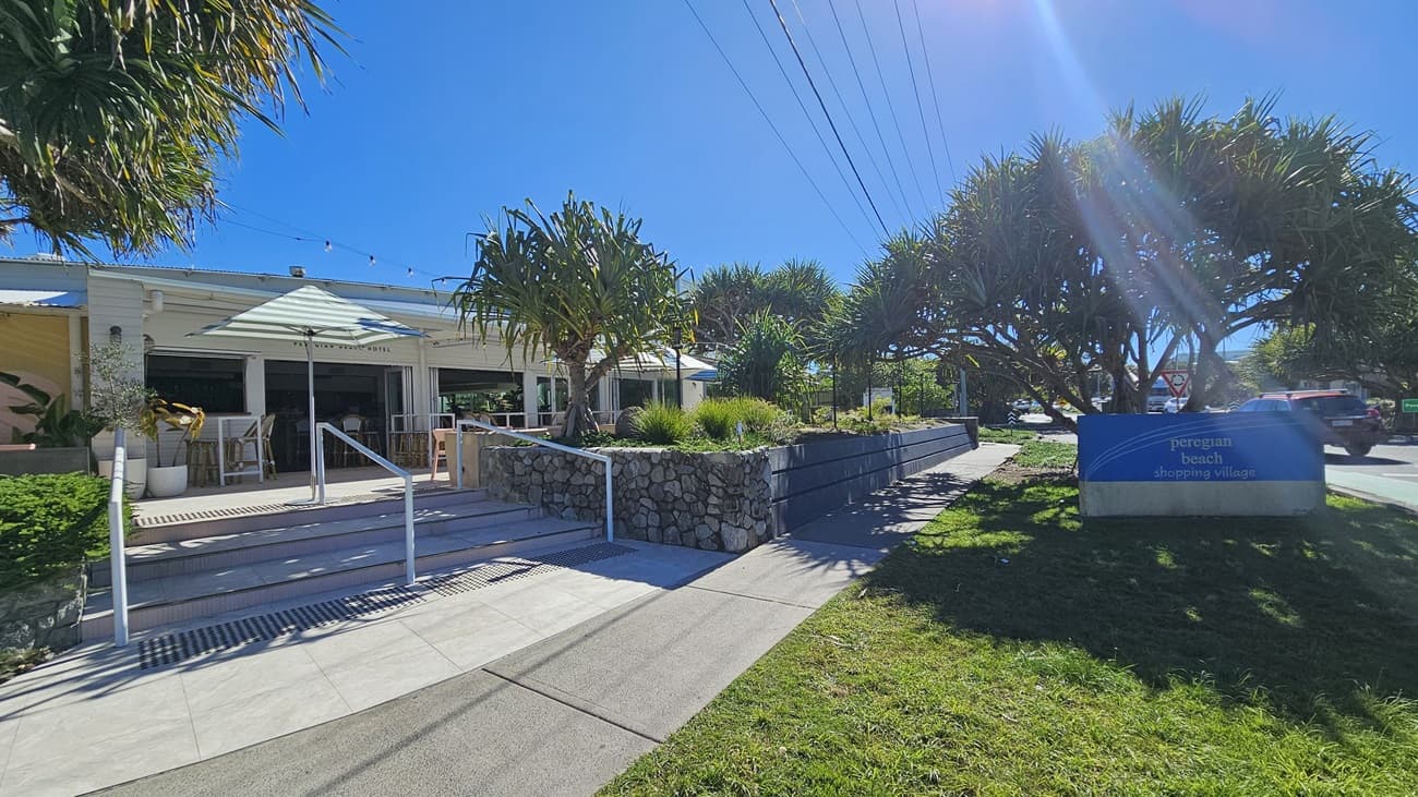 On the left, there is a white building with large windows, a ramp, and steps leading to the entrance. There are white tables and chairs under green umbrellas. In the middle, there is a stone wall with plants on top, separating the building from the pathway. On the right, there is a blue sign for Peregian Beach Hotel next to a grassy area and a road with parked cars. The foreground has a concrete pathway, and the background includes tall trees and power lines.