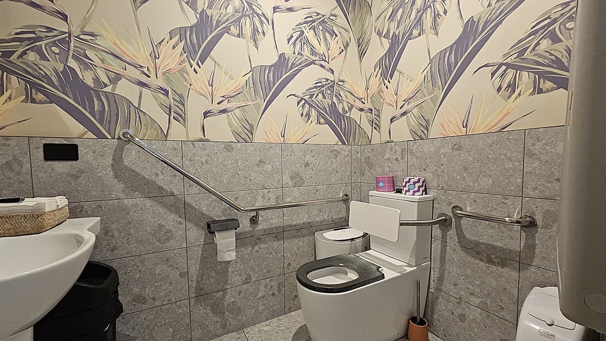 On the left, there is a white door with a wheelchair access sign and a door handle, followed by a wall-mounted baby changing station. On the right, there is a white panelled wall with a black ATM machine. In the middle, there is a long, narrow corridor with a polished concrete floor and white walls. The background features another white door at the end of the corridor. The ceiling is white with recessed lighting and a smoke detector.