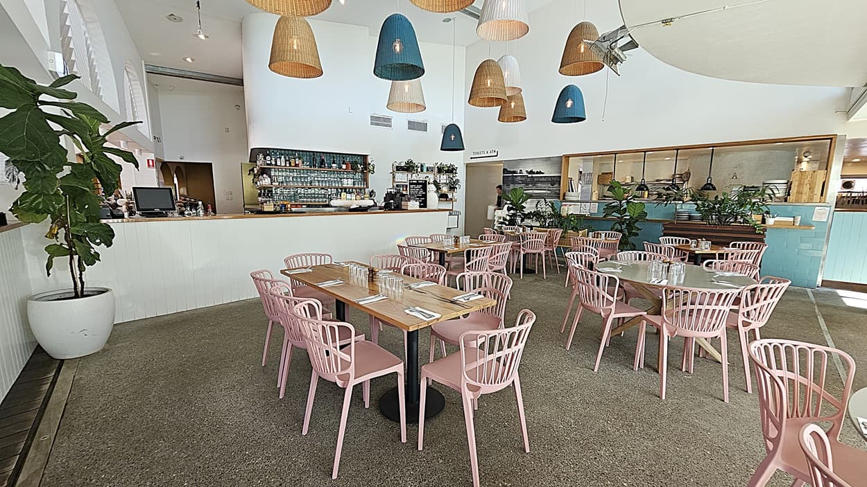 On the left, there is a white counter with a geometric pattern and a potted plant on top. In the middle, there are round tables with pink chairs on a grey carpeted floor, under hanging wicker and blue lamps. The background features a turquoise counter with plants and shelves, and a staircase leading to an upper level. On the right, there is a menu stand on a gold pole, more plants, and a wooden floor section. The ceiling is white with recessed lighting and hanging lamps.