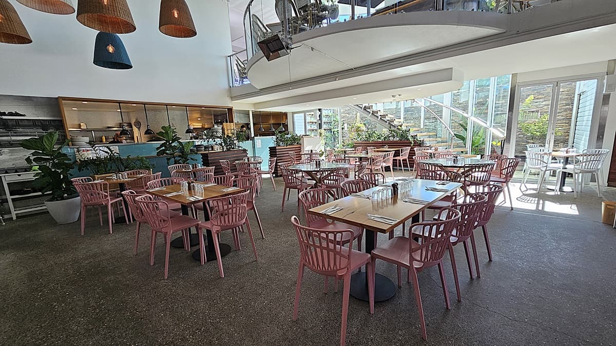 On the left, there are wooden tables with pink chairs, potted plants, and a turquoise counter with shelves behind it. In the middle, there are more wooden tables and pink chairs on a polished concrete floor, with a staircase leading to an upper level with glass railings. The background features large windows and glass doors opening to an outdoor area. On the right, there are additional tables and chairs near the glass doors. The ceiling is white with hanging wicker lamps and recessed lighting.