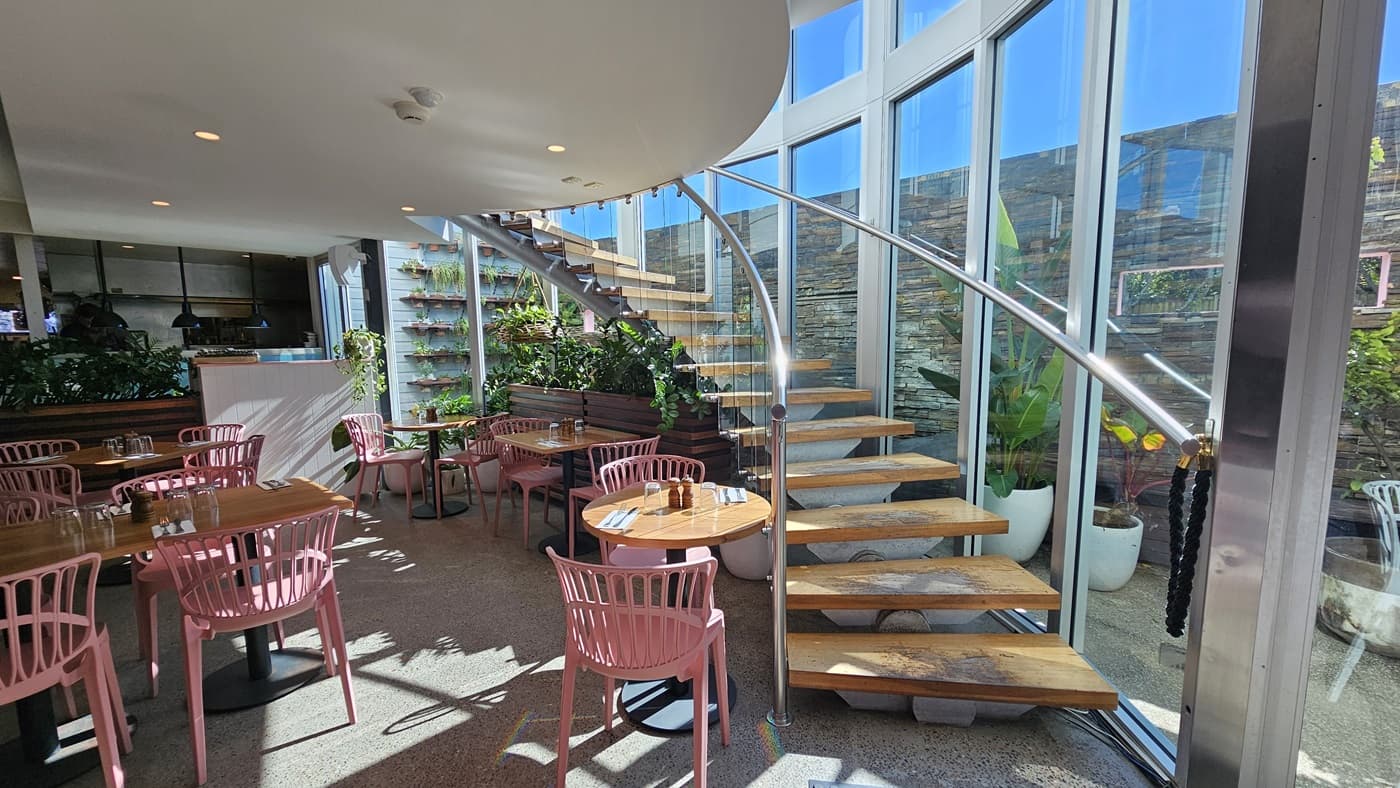 On the left, there are wooden tables with pink chairs on a speckled floor, next to a wooden counter with plants and shelves behind it. In the middle, there is a curved staircase with wooden steps and a glass railing, leading to an upper level. The background features large windows and glass doors, with a stone wall and more plants outside. On the right, there are additional tables and chairs near the windows, with sunlight streaming in. The ceiling is white with recessed lighting.