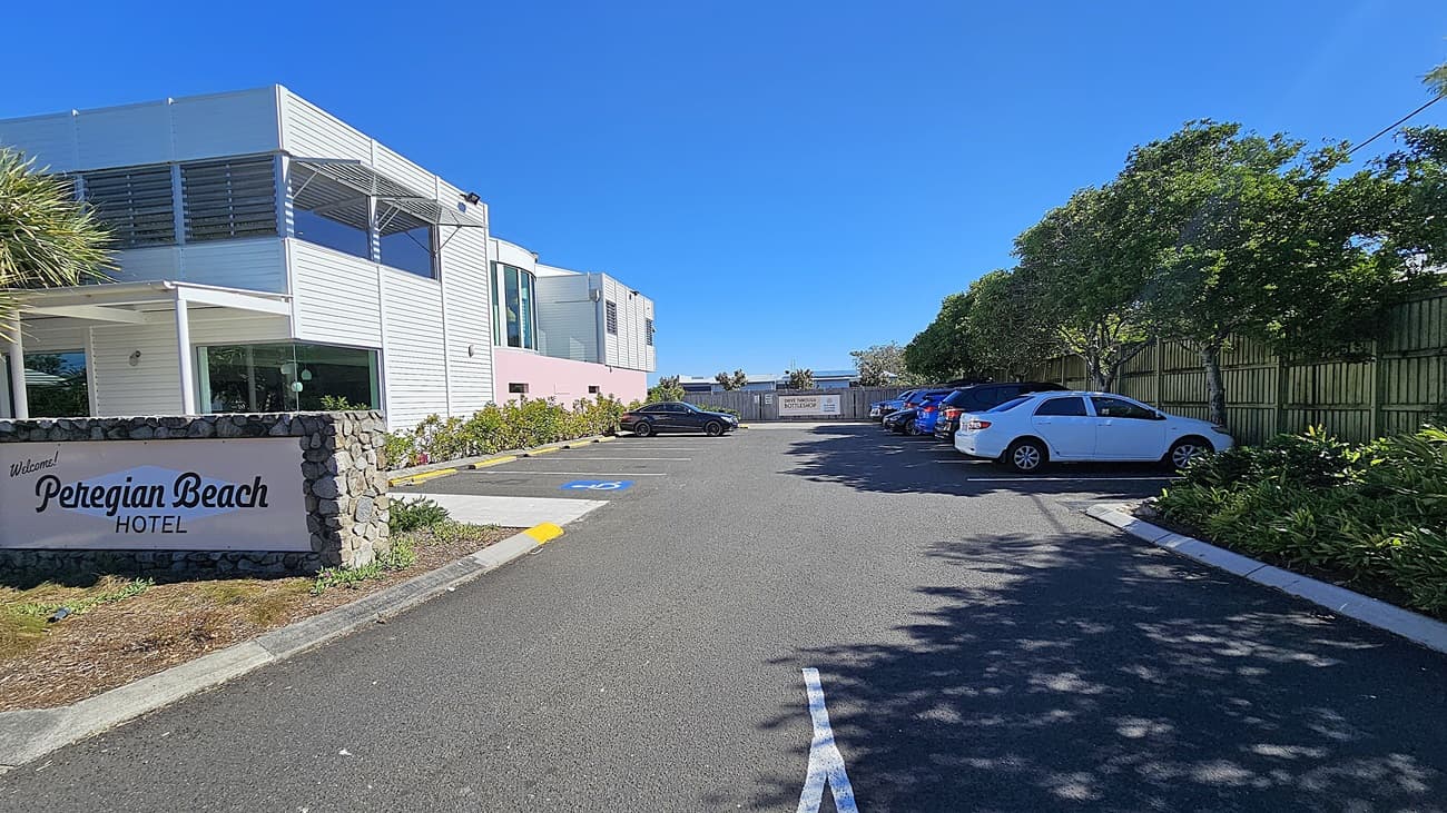 On the left, there is a paved road with a pedestrian crossing and a green bike lane. A white sign reads "Peregian Beach Hotel" next to a landscaped area with trees and shrubs. On the right, there is a large tree with a white lamp post and more landscaped greenery. In the middle, there is a white building with large windows and a pink section, surrounded by a parking area with a few cars.