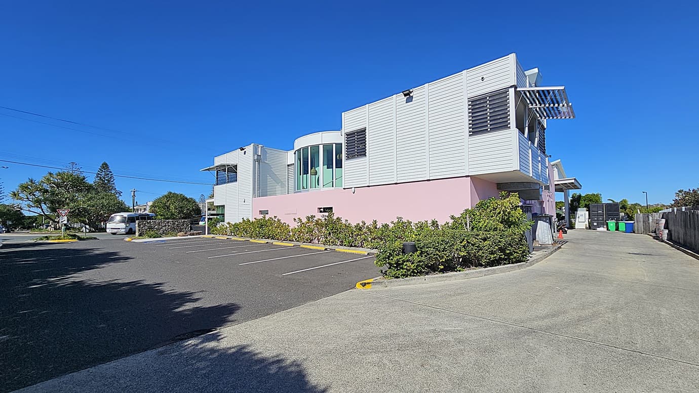 On the left, there is a white building with large windows and a pink lower section, surrounded by landscaped greenery. There are a few parked cars along the side. On the right, there is a driveway leading to a parking area with green, yellow, and blue bins. In the middle, there is an asphalt parking lot with marked spaces and a concrete walkway. The background includes more buildings and trees.