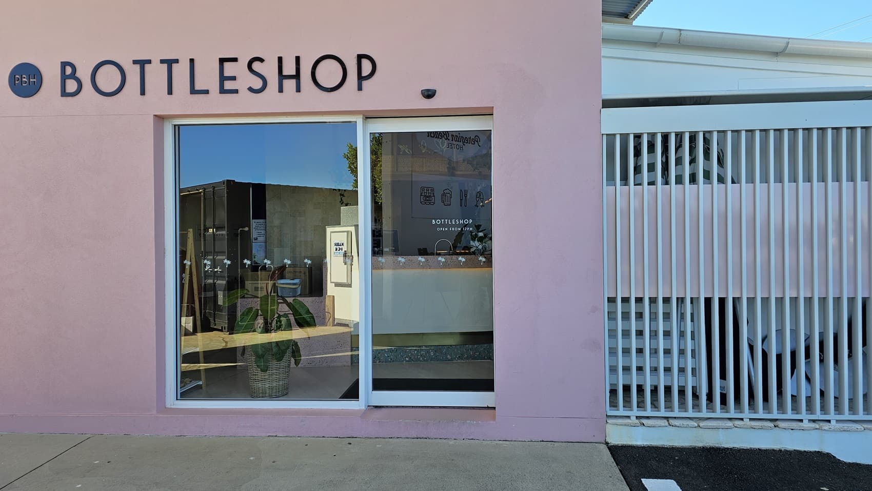 On the left, there is a pink wall with a large window and a glass door, both displaying the word "BOTTLESHOP" above them. Inside the window, there are shelves with bottles and a potted plant. On the right, there is a white slatted fence with a staircase behind it. The floor is a mix of concrete and asphalt. The ceiling is not visible.