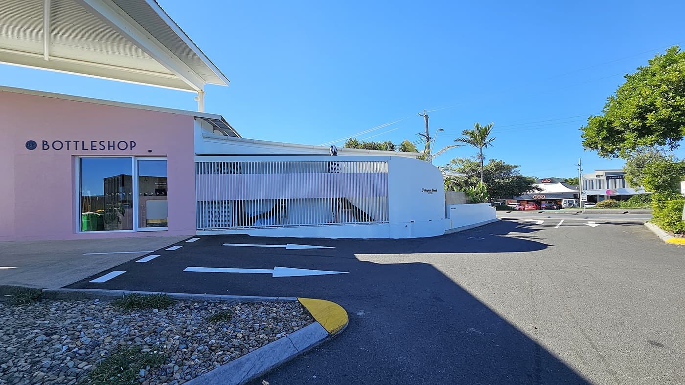On the left, there is a pink building with a large window and a glass door, both labelled "BOTTLESHOP." Inside the window, there are shelves with bottles and a green bin. On the right, there is a white slatted fence with a ramp behind it, leading to a parking area. In the background, there are more buildings, trees, and a few parked cars. The floor is asphalt with white directional arrows and a section of gravel with plants.