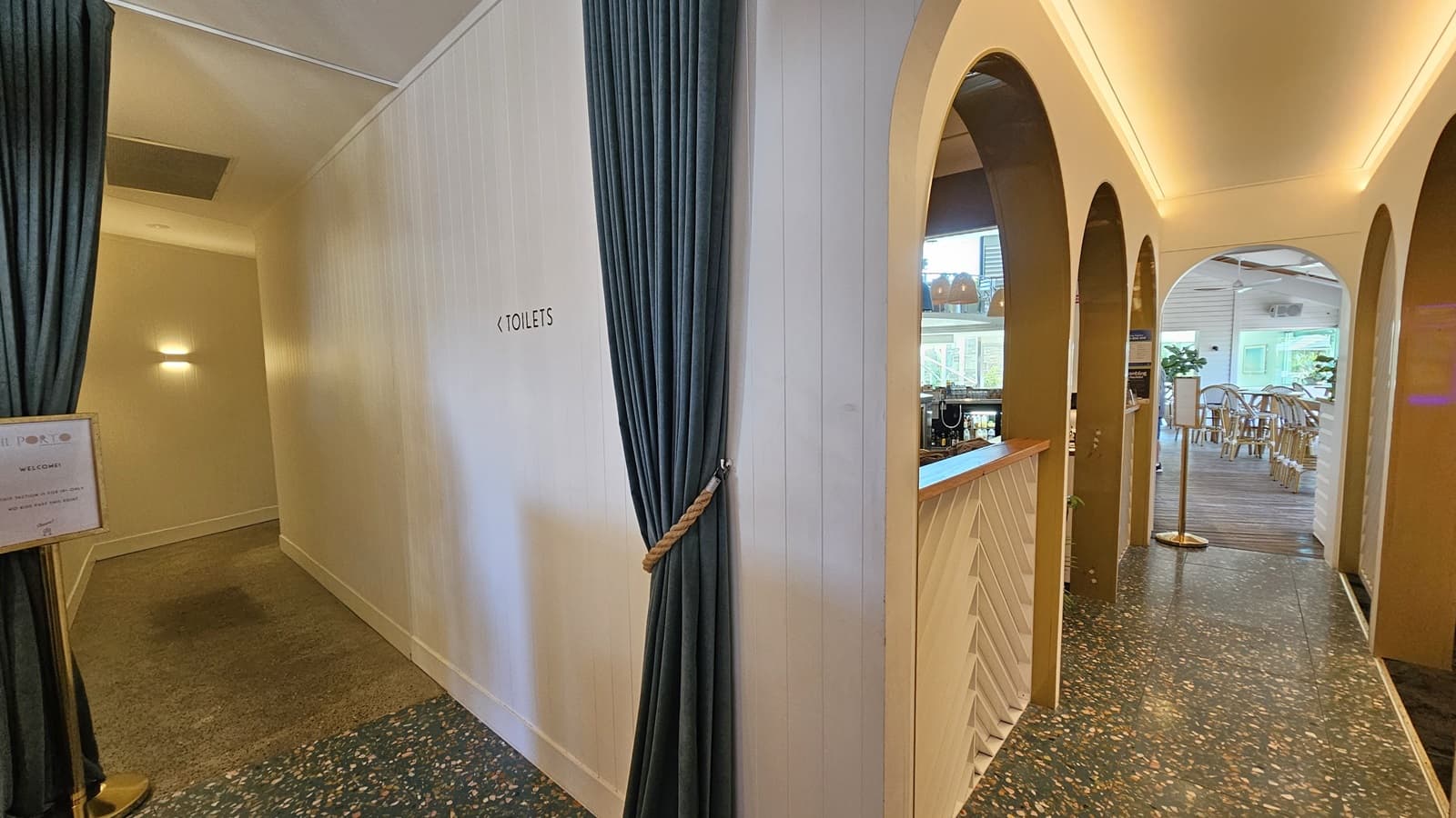 On the left, there is a hallway with a dark green curtain tied back, leading to the toilets, with a sign on the wall. The floor is carpeted in grey. In the middle, there is a white wall with a counter featuring a white front and wooden top, with shelves holding various items above it. On the right, there are arched doorways leading to a dining area with wooden tables and chairs on a speckled floor. The ceiling has recessed lighting and wooden beams.
