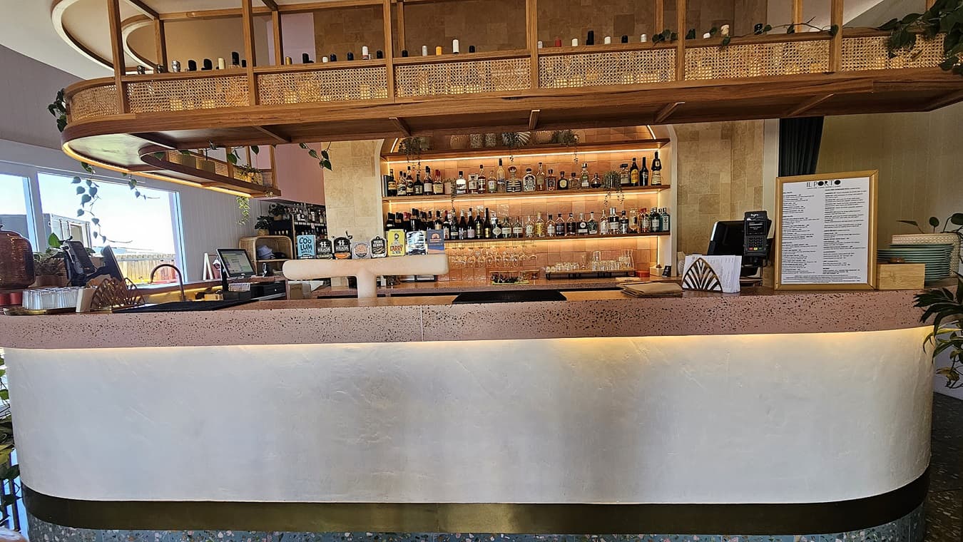 On the left, there are plants and decorative items on a counter, with a glimpse of outdoor seating through a window. In the middle, there is a curved bar counter with a pink terrazzo top and a white front, displaying various bottles and a menu. The background features shelves filled with bottles and glasses, with a wooden wall behind them. On the right, there is a menu stand and more plants. The floor is speckled, and the ceiling has wooden panels with recessed lighting and hanging shelves.