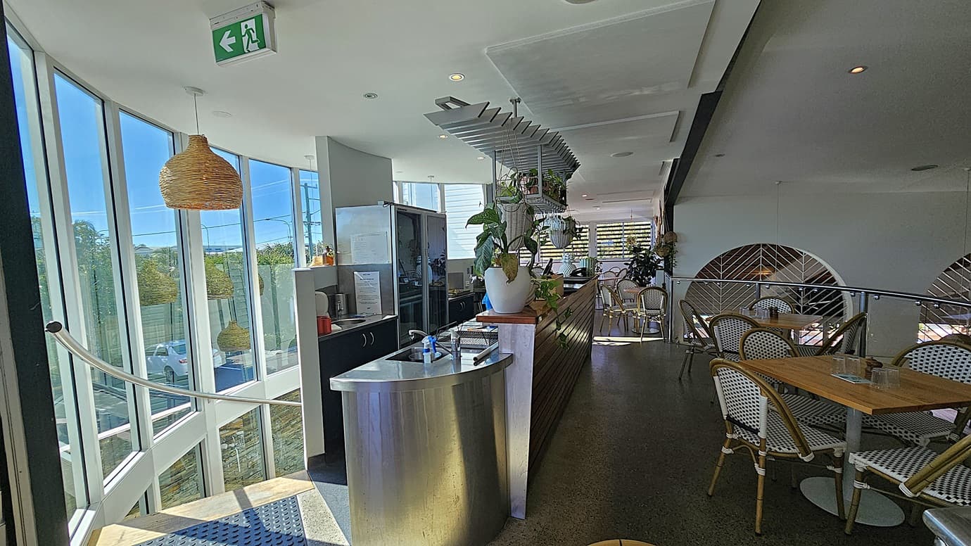 On the left, there is a curved staircase with metal railings leading down, next to large windows. In the middle, there is a stainless steel counter with a potted plant and a coffee machine, with hanging shelves above. The background features more windows and a view of trees outside. On the right, there are wooden tables with wicker chairs, and a partition with a geometric design. The floor is polished concrete, and the ceiling is white with recessed lighting and a hanging wicker lamp.