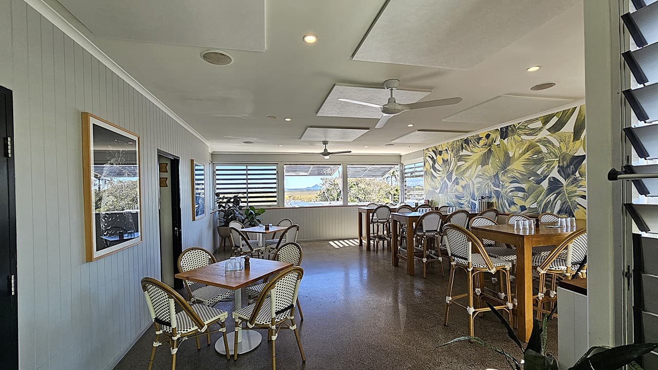 On the left, there are square wooden tables with white and blue woven chairs, next to a wall with framed pictures. In the middle, there are more tables and chairs on a polished concrete floor, leading to large windows with louvres and a view of trees outside. The background features a wall with a green and yellow leaf pattern. On the right, there are additional wooden tables and chairs. The ceiling is white with recessed lighting, ceiling fans, and rectangular acoustic panels.