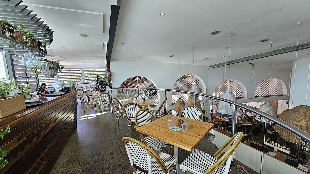 On the left, there is a wooden counter with plants and hanging glasses above it, next to a row of windows with louvres. In the middle, there are square wooden tables with white and blue woven chairs on a polished concrete floor, bordered by a glass railing overlooking the lower level. The background features large arched windows with a chevron pattern. On the right, there are more tables and chairs near the railing. The ceiling is white with recessed lighting and hanging wicker lamps.