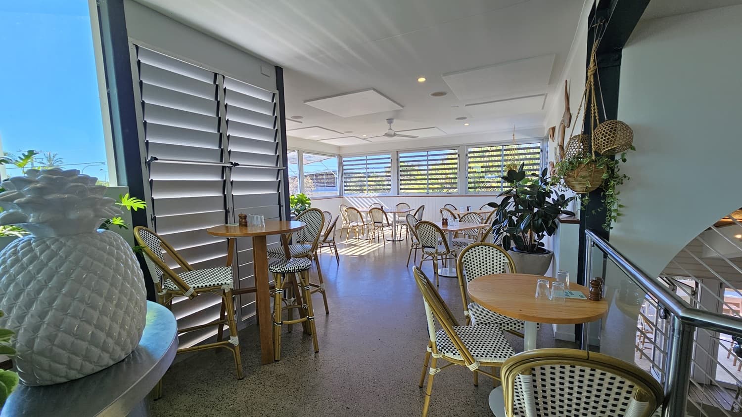 On the left, there is a tall wooden table with two woven chairs and a large white pineapple decoration on a metal counter. In the middle, there are more wooden tables and woven chairs on a polished concrete floor, with large windows featuring louvres allowing light in. The background has additional tables and chairs near the windows. On the right, there is a glass railing with hanging wicker baskets and plants. The ceiling is white with recessed lighting and ceiling fans.