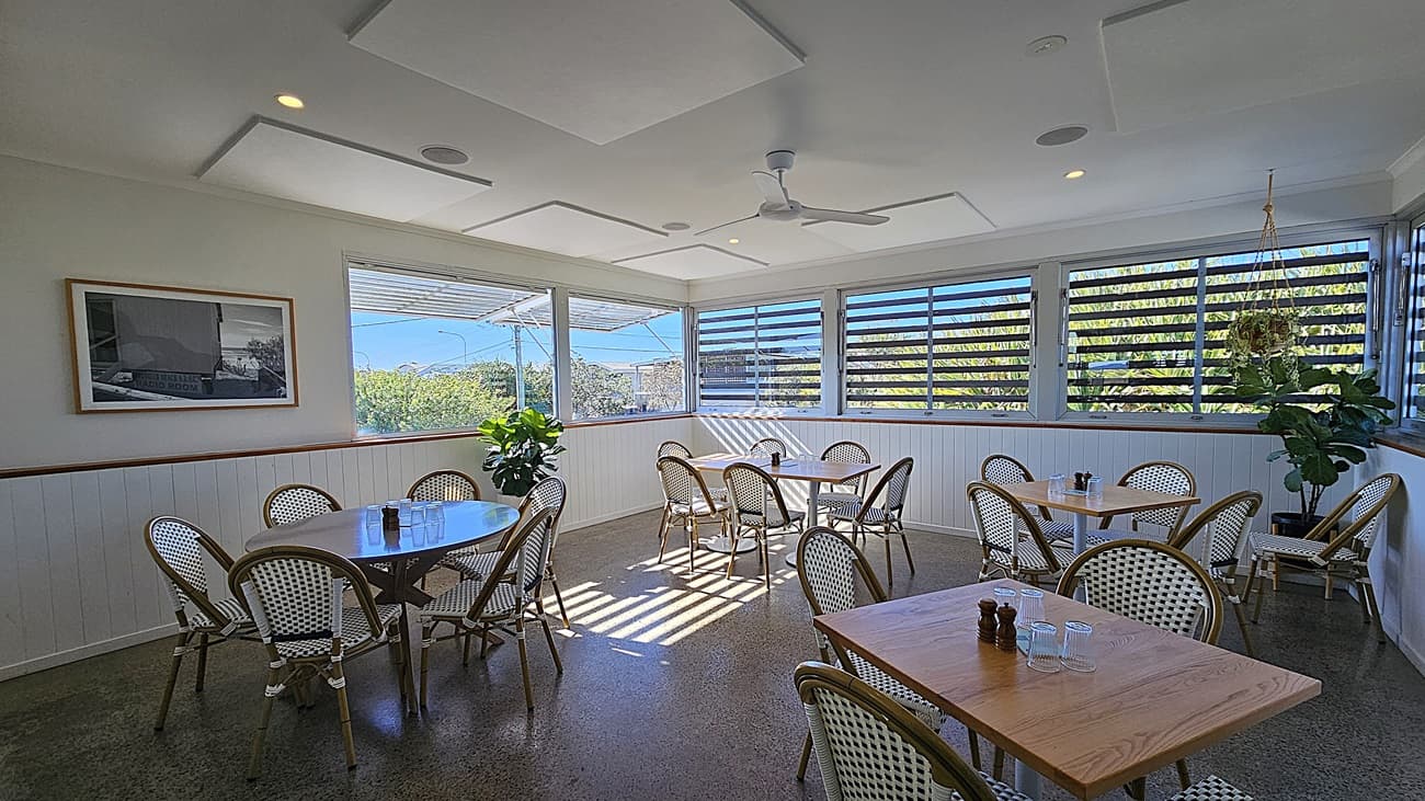 On the left, there are wooden tables with white and green woven chairs, next to a white wall. In the middle, there are more tables and chairs on a polished concrete floor, with sunlight streaming through louvred windows. The background features large windows with louvres and a view of greenery outside. On the right, there are additional tables and chairs, along with potted plants near the windows. The ceiling is white with recessed lighting, ceiling fans, and  acoustic panels.