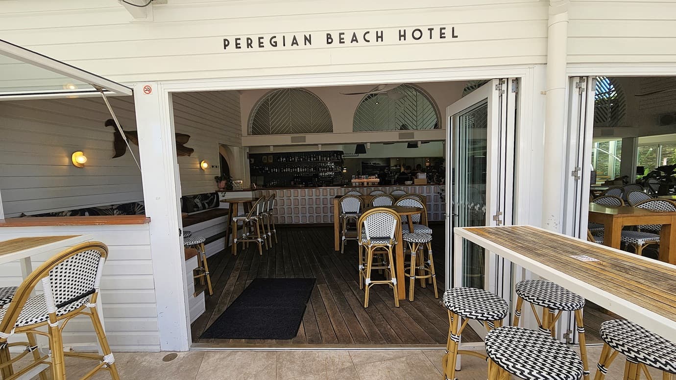 On the left, there is the entrance to the Peregian Beach Hotel with large open sliding doors, revealing a bar area with high stools and tables inside. In the middle, there are high tables with wicker stools on a tiled floor, under a white pergola ceiling with string lights. On the right, there are more tables and chairs, some under large umbrellas, with a mix of pink and brown seating. The background includes green plants and trees, with a glimpse of a road and more seating.
