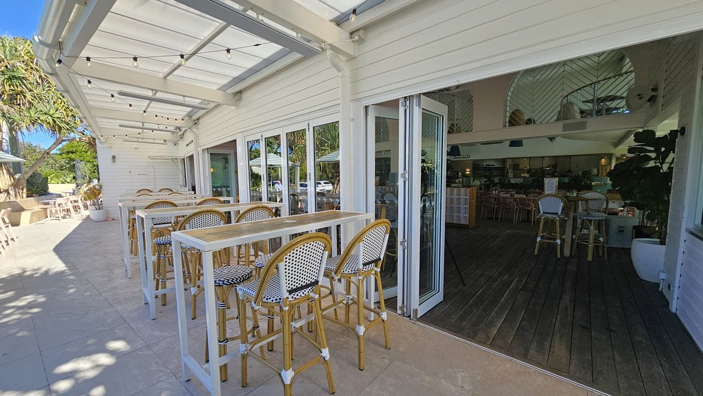 On the left, there are high white tables with wicker stools on a tiled floor, next to a white wall with large windows. In the middle, the entrance to the Peregian Beach Hotel has open sliding glass doors leading to a bar area with more high stools and tables on a wooden floor. On the right, there are additional high tables and stools inside the bar area. The ceiling is a white pergola with string lights hanging above the outdoor seating area. The background includes more seating and greenery.