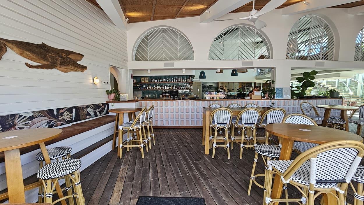 On the left, there is a wooden bench with a patterned cushion and a small wooden table, next to a white wall with a wooden fish decoration. In the middle, there is a bar counter with a patterned front, high stools, and a variety of bottles and glasses behind it. The floor is wooden. On the right, there are more tables and wicker chairs with patterned cushions. The ceiling has wooden panels and white beams, with ceiling fans and lights. The background includes arched windows and more seating.