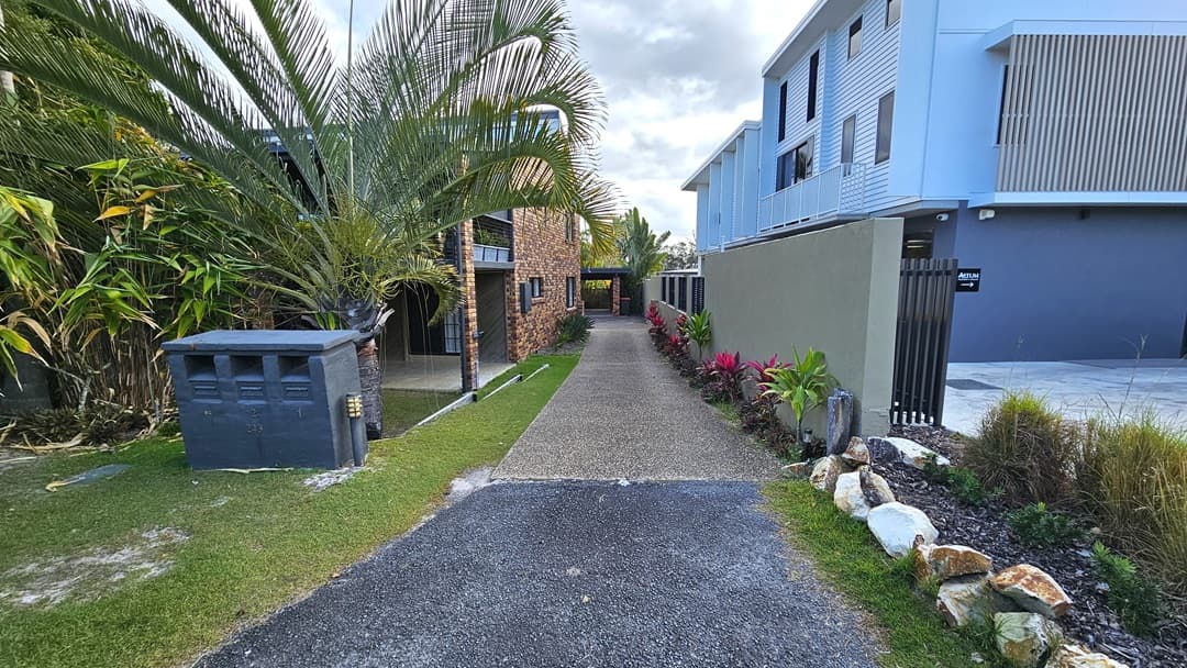 On the left, there is a paved footpath and a road with a green utility pole. In the middle, there is a brick house with large windows and a driveway leading to a garage. On the right, there are tall palm trees and dense greenery surrounding the house. The foreground features a mix of grass and a paved driveway.