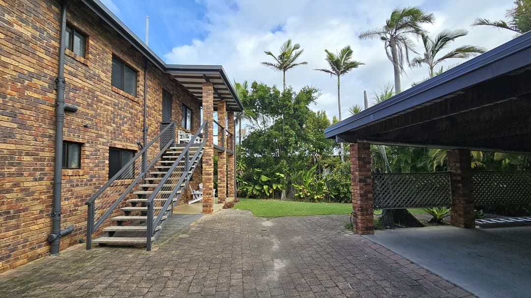 On the left, there is a two-storey brick house with an external staircase leading to the upper floor and a small balcony with white chairs. In the middle, there is a paved area extending towards a grassy lawn with tall plants and trees in the background. On the right, there is a covered carport with a brick structure and a lattice fence. The floor is a combination of paving stones and concrete. The ceiling of the carport is wooden with exposed beams.