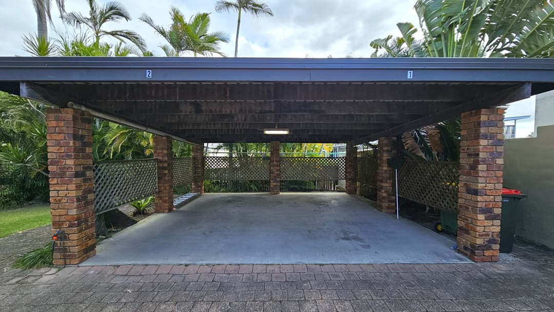 On the left, there is a brick pillar supporting the carport roof, with a lattice fence and lush green plants behind it. In the middle, there is an open concrete floor space under the carport, with a light fixture on the wooden ceiling. On the right, there is another brick pillar, a lattice fence, and more green plants. The floor is a mix of concrete under the carport and paving stones in the foreground. The ceiling is wooden with exposed beams.