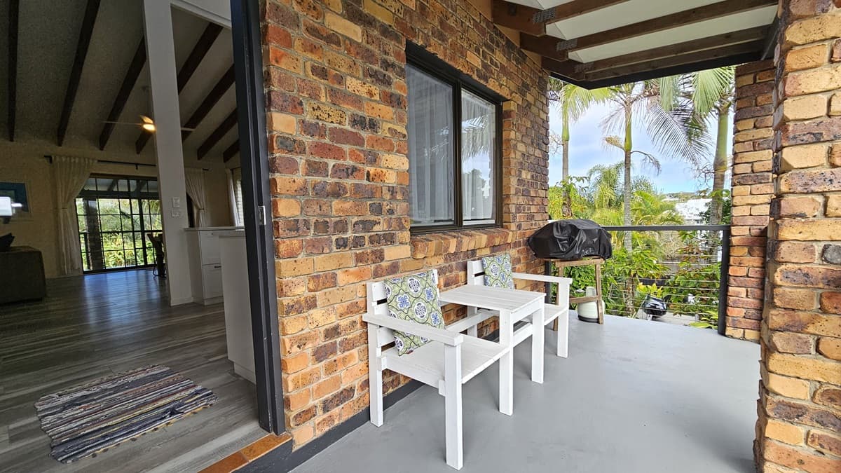 On the left, there is an interior space with wooden beams on the ceiling, a rug on the wooden floor, and a view of a living area with large windows. In the middle, there is a brick wall with a window and a small balcony area. On the right, the balcony has a white wooden bench with cushions, a barbecue grill, and potted plants. The floor of the balcony is concrete, and the ceiling has exposed wooden beams.
