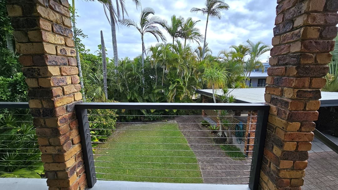 On the left, there is a brick pillar with a view of dense green foliage and tall palm trees. In the middle, there is a balcony with a metal railing and cable wires, overlooking a grassy lawn and a paved pathway. On the right, there is another brick pillar, more palm trees, and part of a building with a metal roof. The floor of the balcony is concrete.