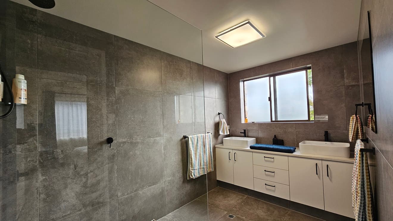 On the left, there is a bathroom with a large glass-enclosed shower featuring dark grey tiles and a towel hanging on a rail. In the middle, there is a white wall with a partially open door leading to another bathroom. On the right, the second bathroom has white brick walls, a small window, a round mirror, a sink, and a toilet. The floor in both bathrooms is covered with dark grey tiles, and the ceiling is white with recessed lighting.