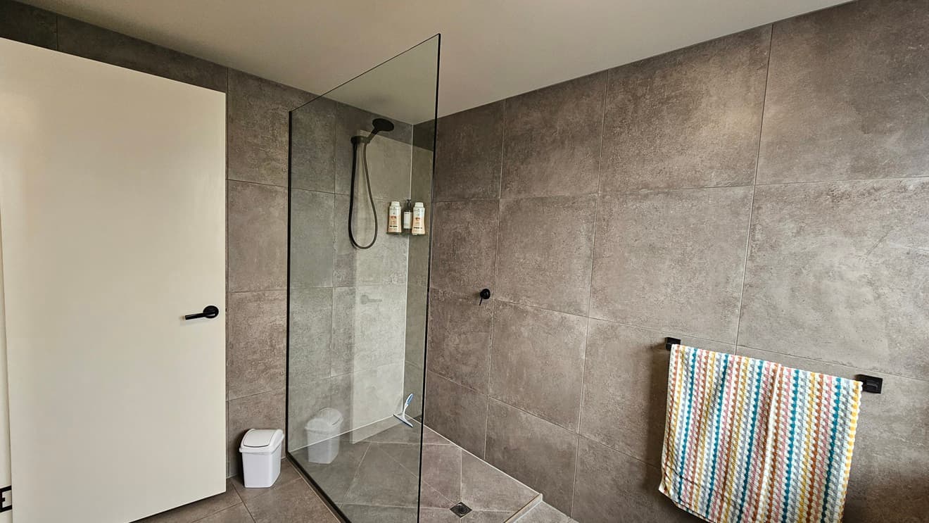 On the left, there is a white door with a black handle and a small white bin on the floor. In the middle, there is a large glass-enclosed shower with dark grey tiles, a black showerhead, and bottles on a built-in shelf. On the right, there is a wall with dark grey tiles and a colourful striped towel hanging on a black rail. The floor is covered with matching dark grey tiles, and the ceiling is white.