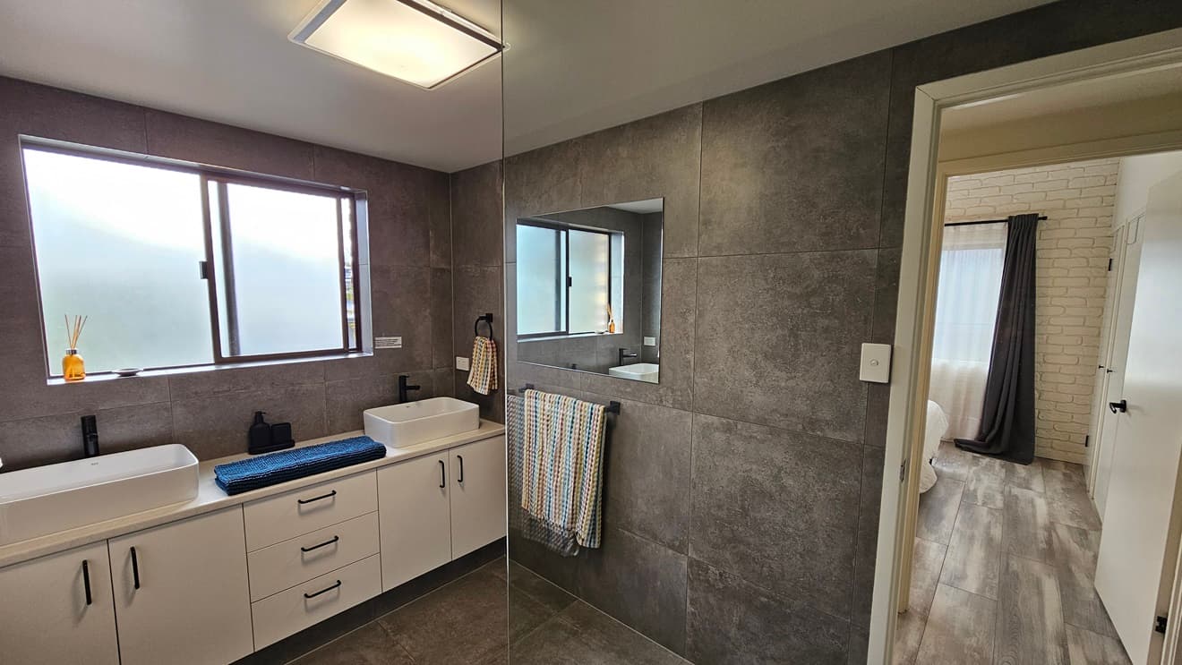 On the left, there is a double sink vanity with white cabinets, black handles, and two white basins. Above the vanity, there is a large window with frosted glass. In the middle, there is a wall with dark grey tiles and a mirror. On the right, there is a doorway leading to a room with a bed and a large window with sheer curtains. The floor is covered with dark grey tiles in the bathroom and wooden laminate in the adjacent room. The ceiling is white with a rectangular recessed light.