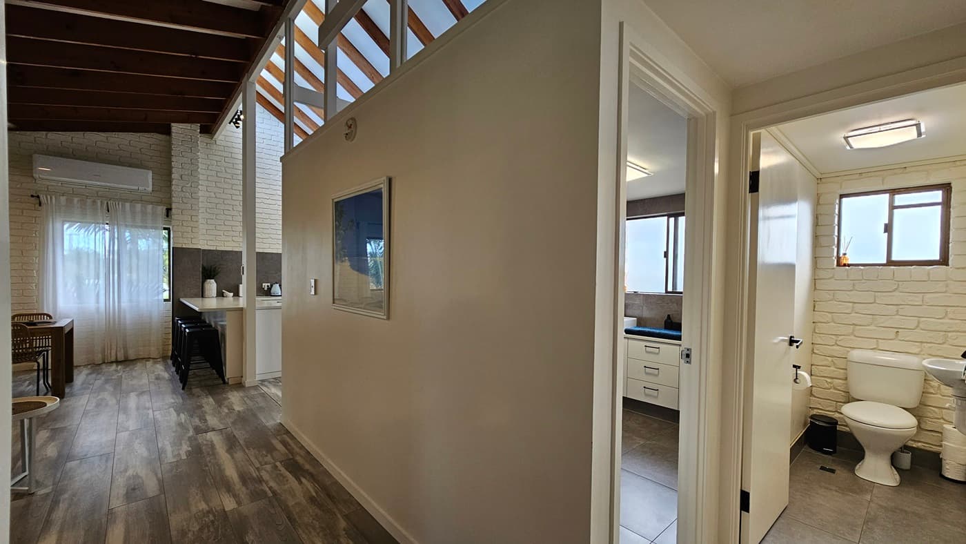 On the left, there is a dining area with a wooden table and chairs, a white brick wall, and a large window with sheer curtains. In the middle, there is a partial wall with a framed picture and a doorway leading to a bathroom. On the right, the bathroom has white brick walls, a window with frosted glass, a white vanity with a sink, and a toilet. The floor is covered with dark wood laminate, and the ceiling has exposed wooden beams and a skylight.